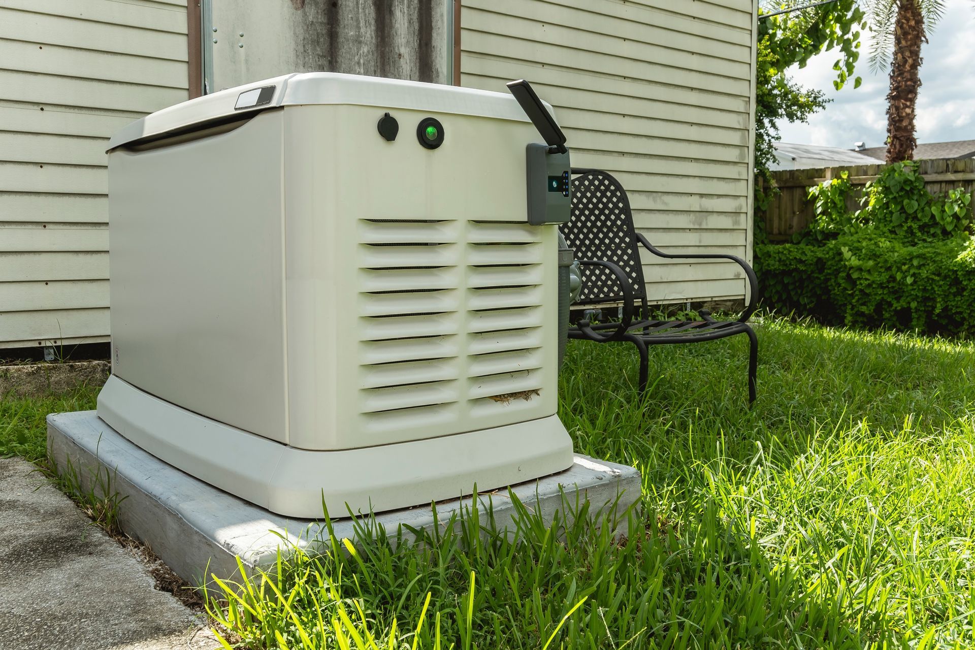 A beige home generator sits on a concrete pad in a grassy yard, next to a house.