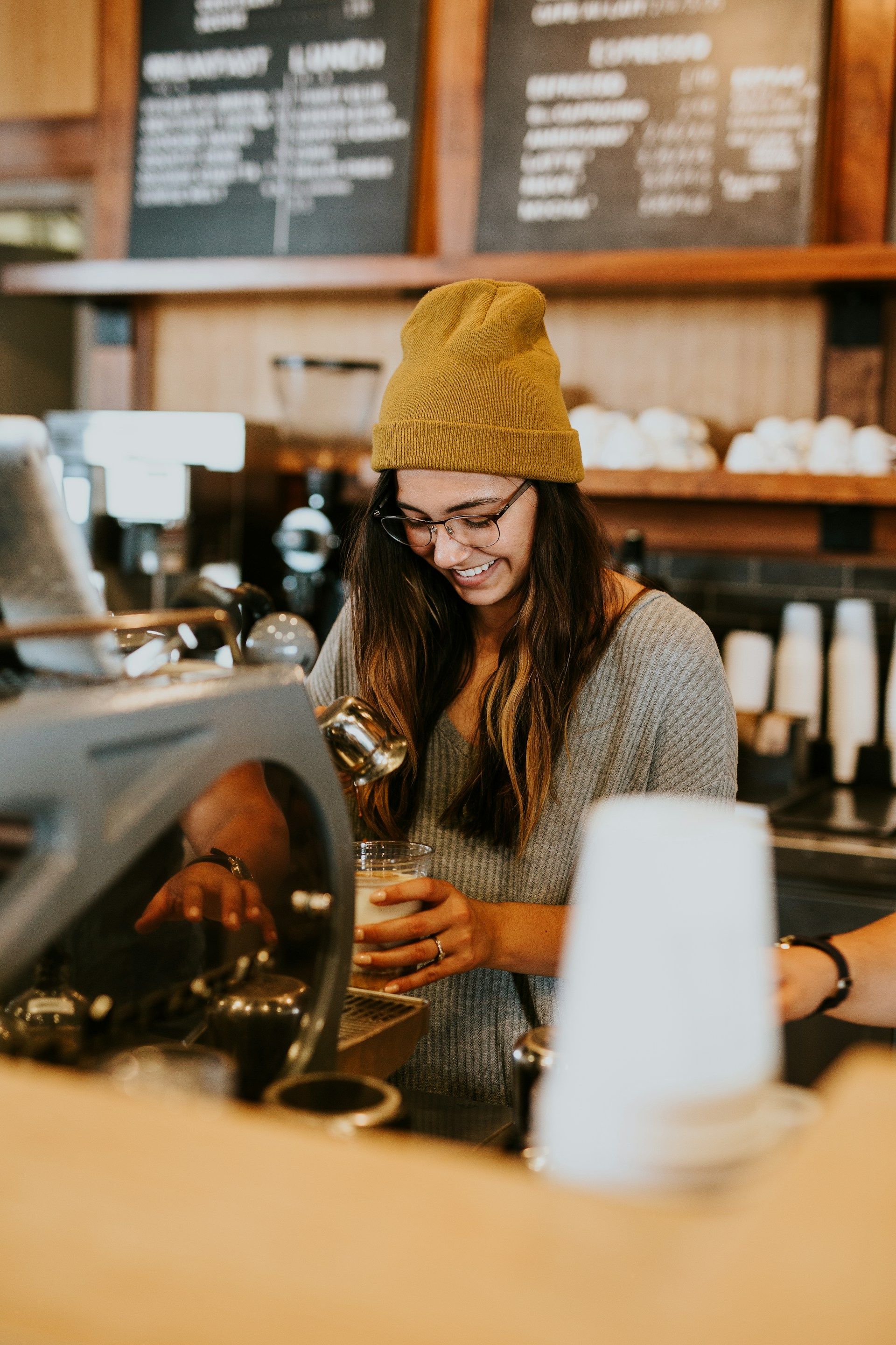 Barista pouring coffee in a cozy café, wearing a mustard knit beanie and glasses.