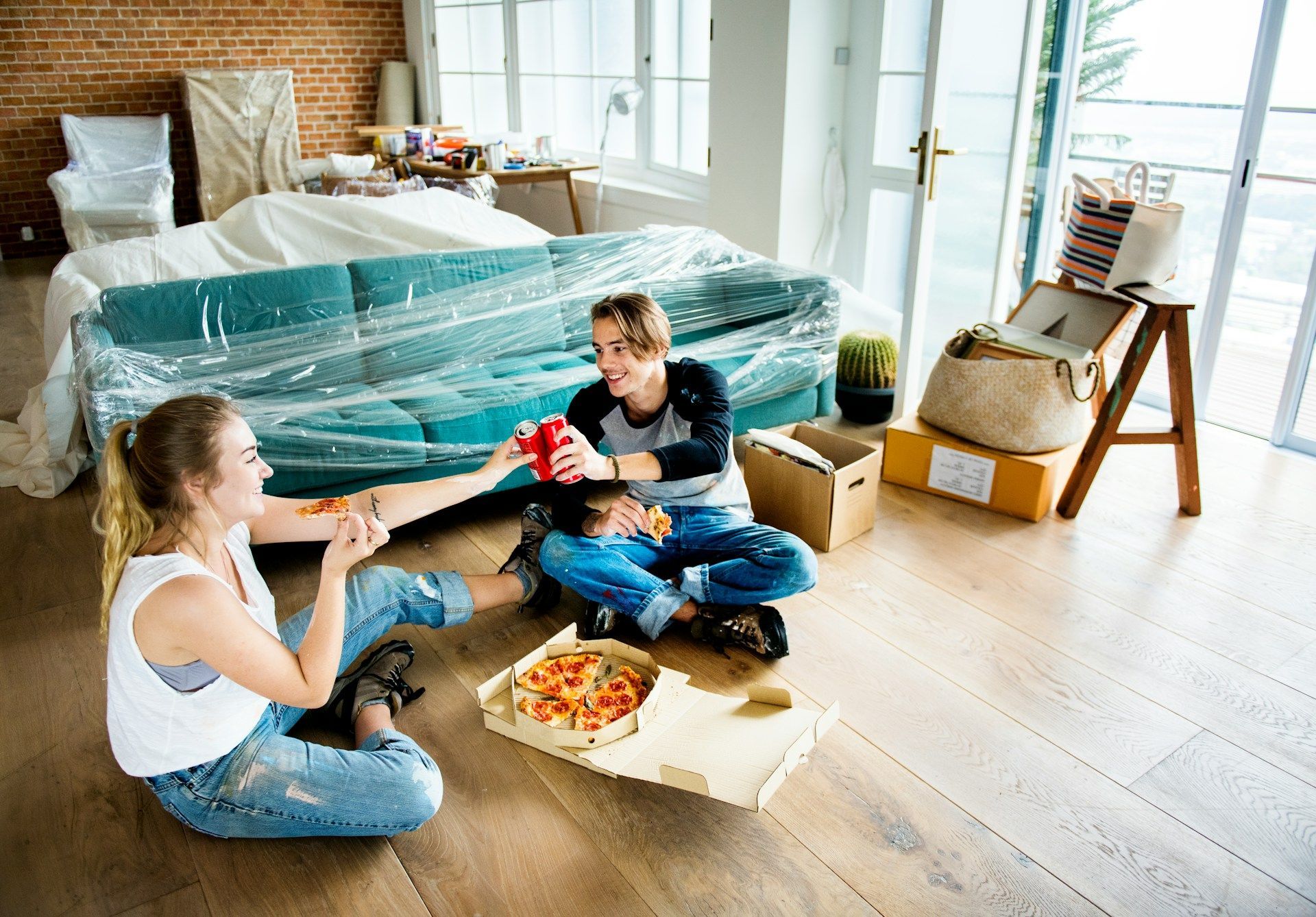 Two children sitting on a living room floor sharing pizza near a wrapped couch and moving boxes