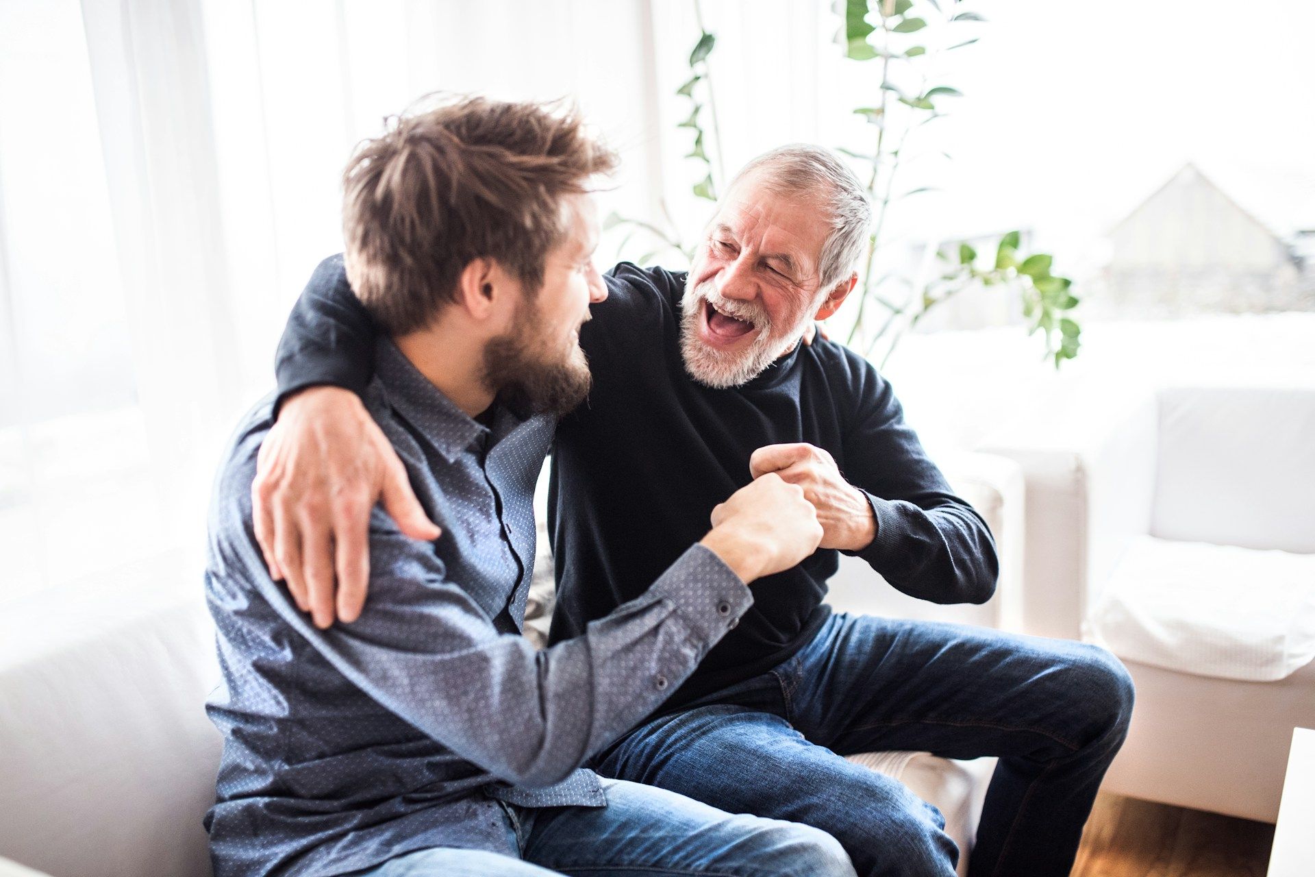 Two men sit on a couch, talking warmly and holding each other’s hands in a bright living room.