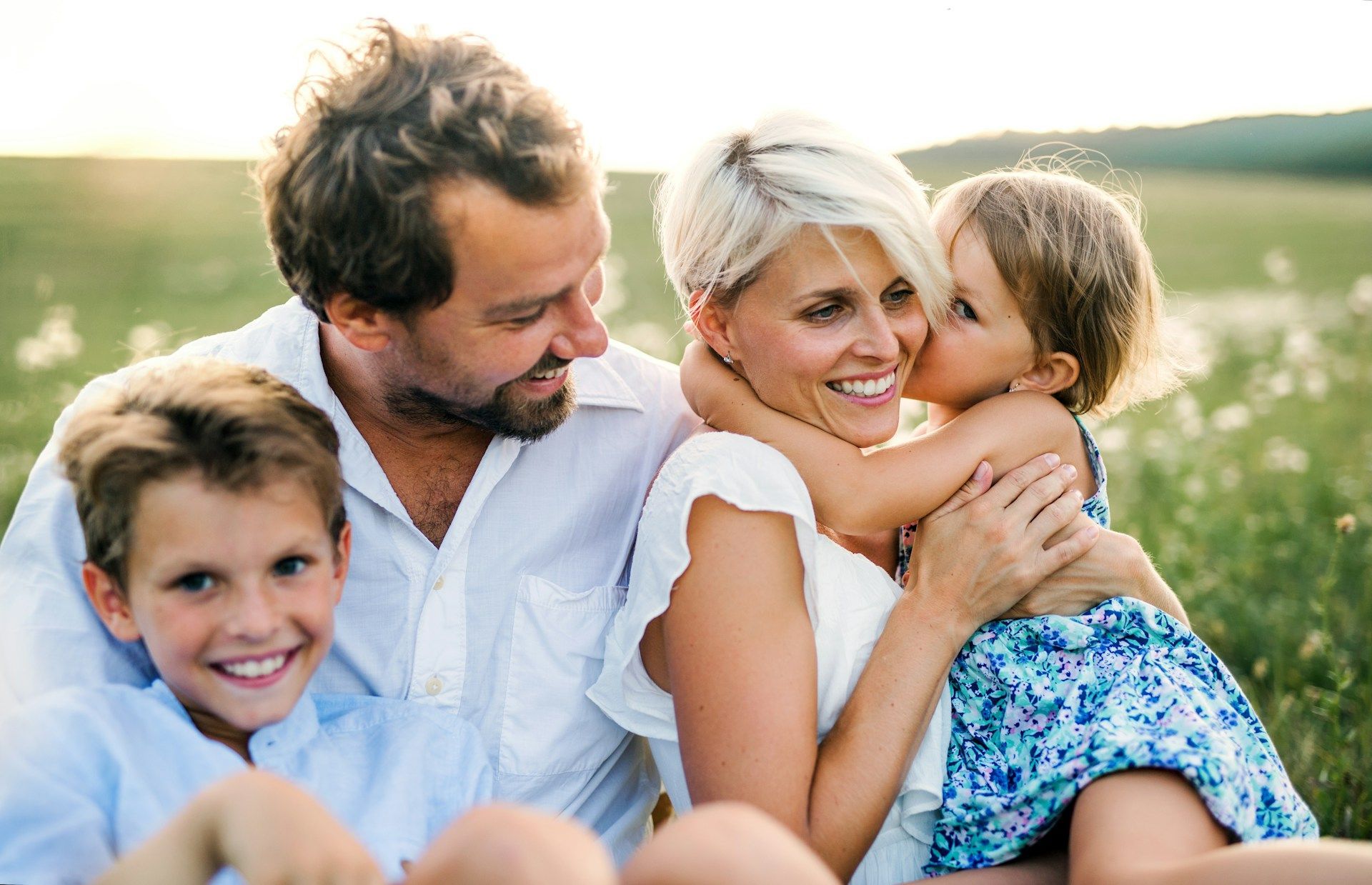 A smiling family of four sitting outdoors in a sunny field, with two children hugging their parents.