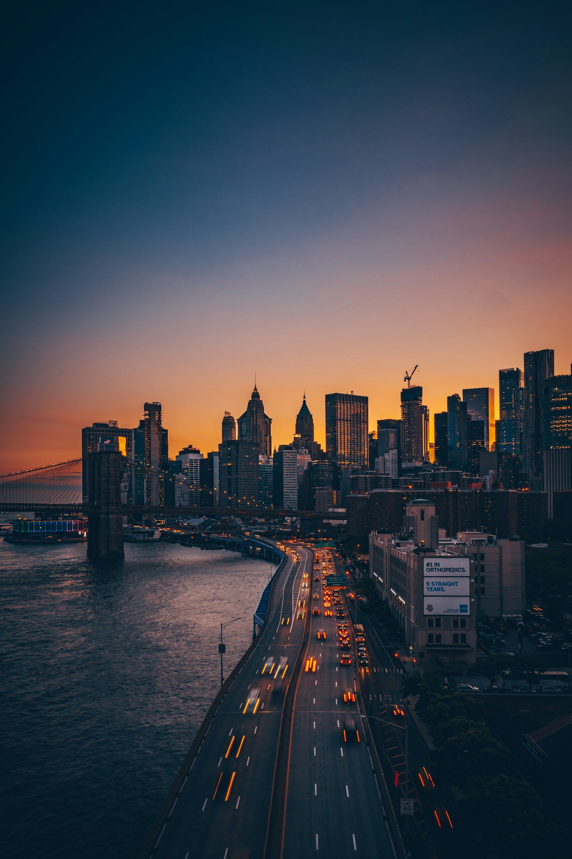 City skyline at dusk with lit traffic on a bridge over water under an orange-blue sunset sky