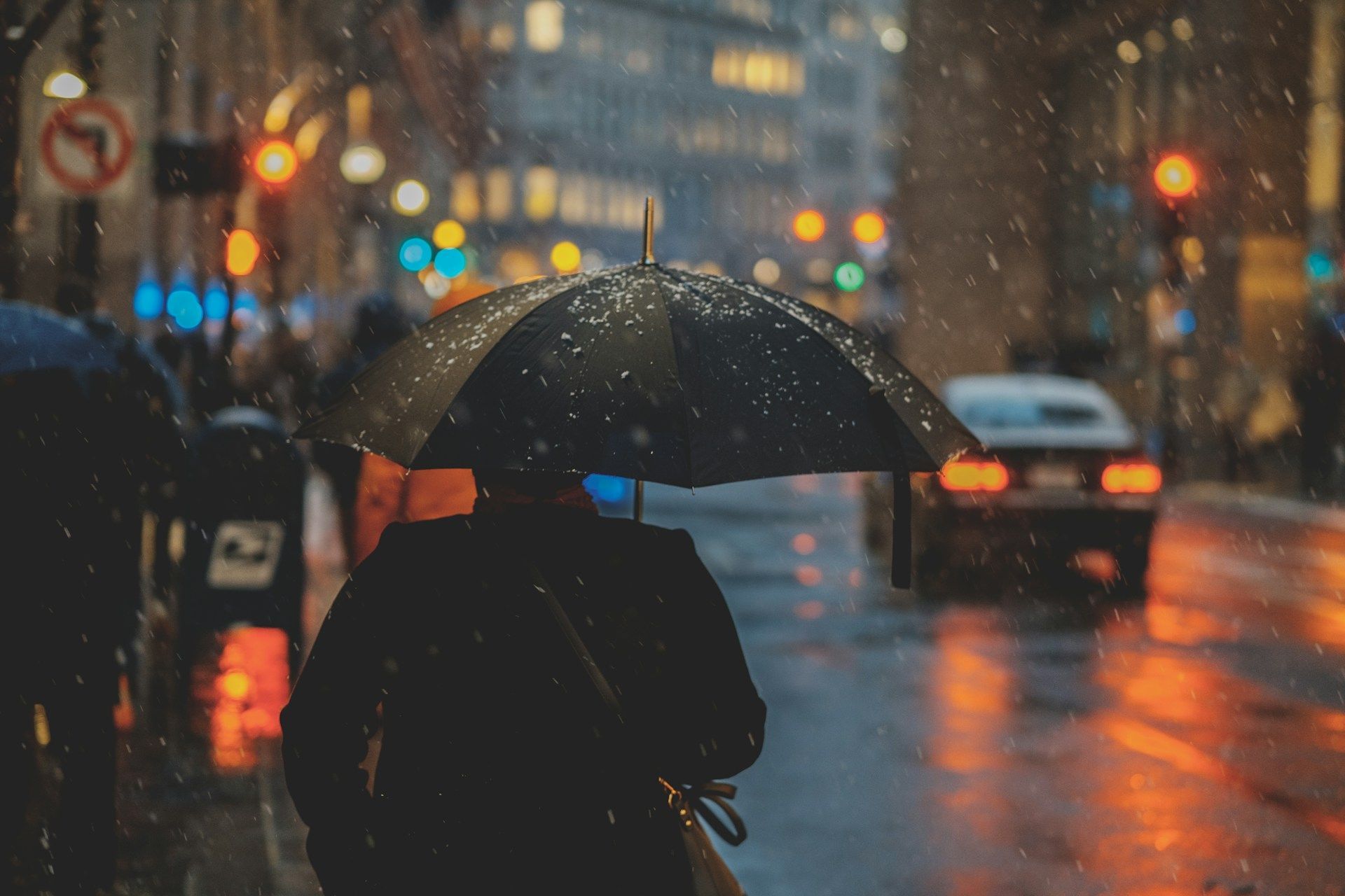 Person with black umbrella walking on a rainy city street at night, with blurred car lights and reflections.