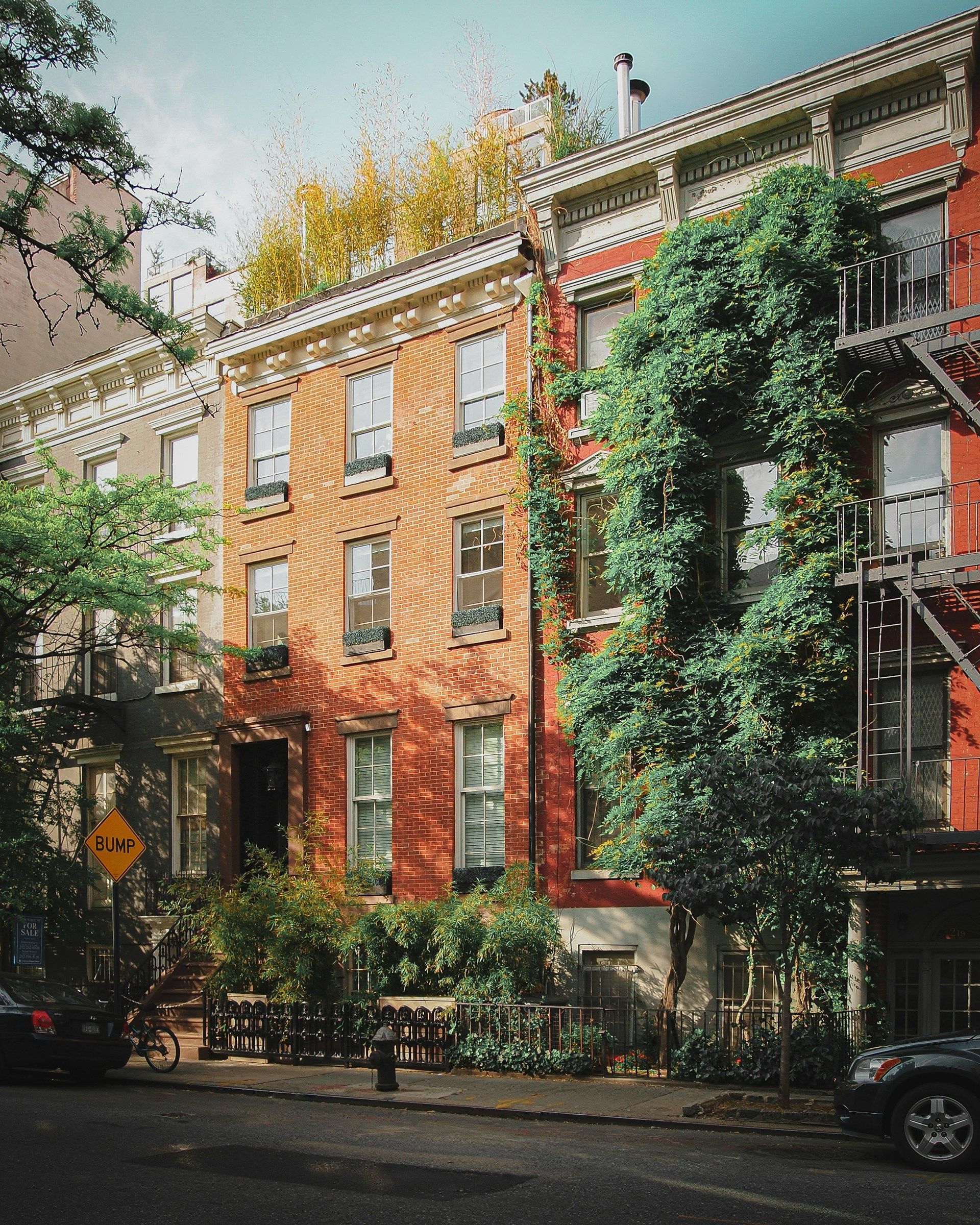 Brick townhouse street with tall green ivy, fire escapes, and parked cars on a quiet city block