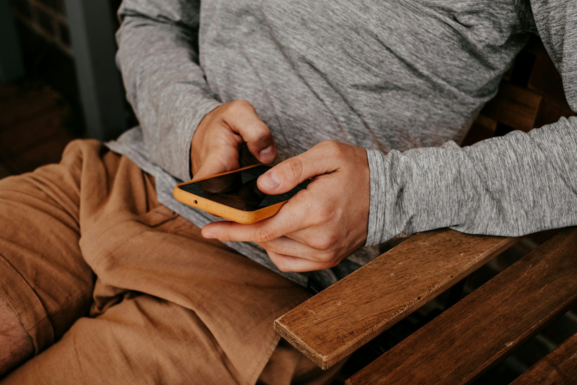 Person using a smartphone while seated in a brown chair, wearing a gray top and tan pants