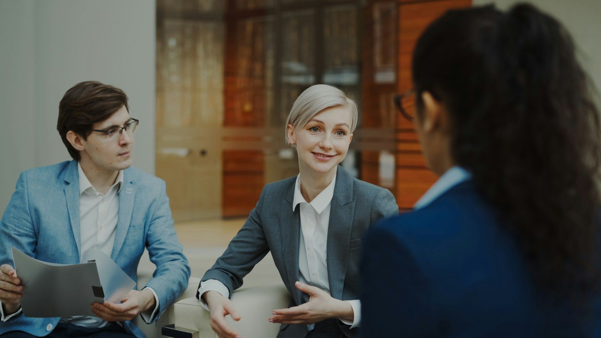 Three people in business attire discuss documents in a modern office conference room.