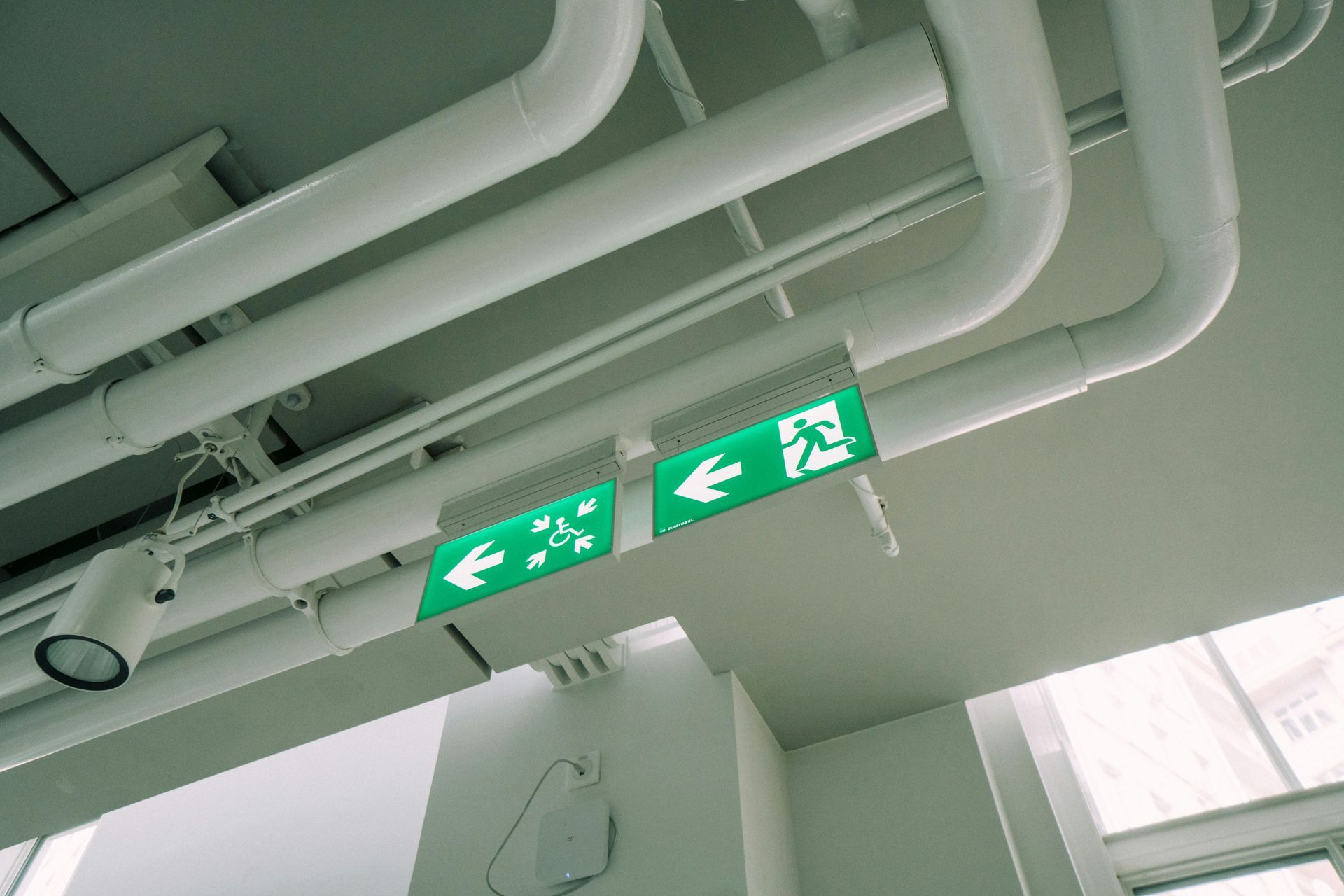 Green emergency exit signs with left arrows on a ceiling near exposed pipes.