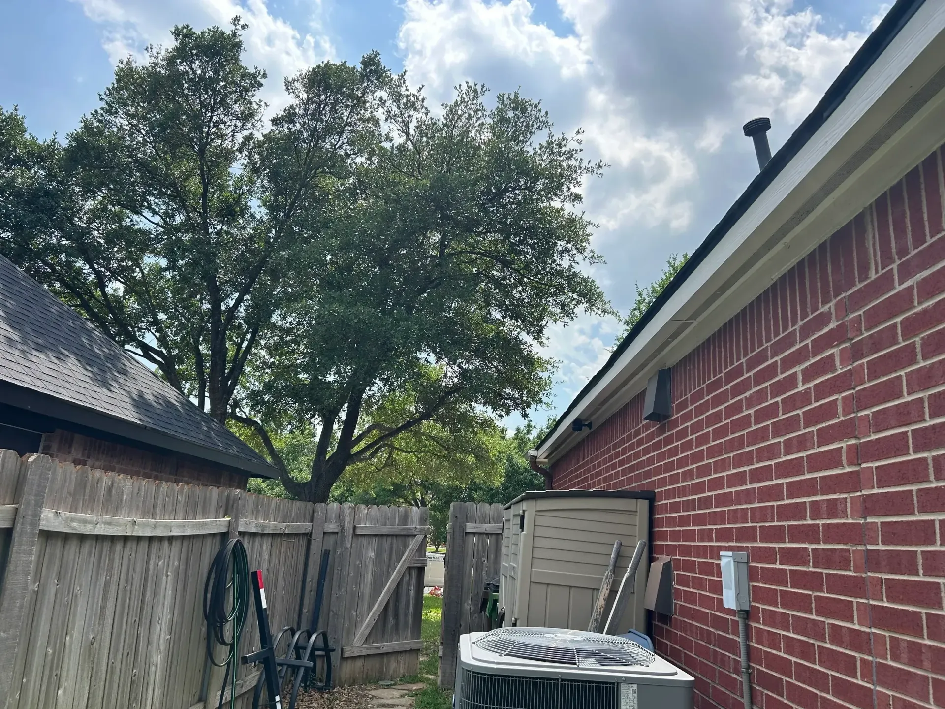 Backyard view with a brick wall, shed, air conditioner, wooden fence, and trees under a cloudy sky.