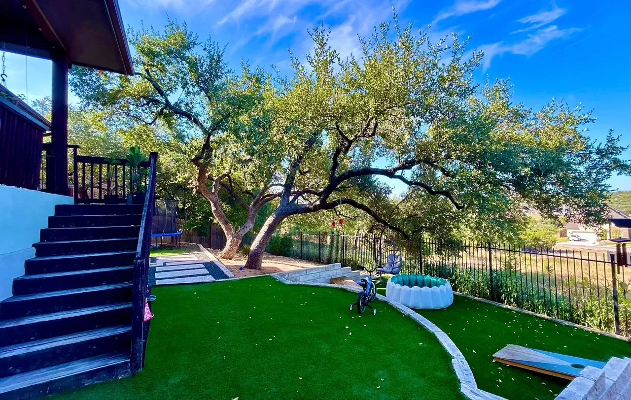 A backyard with a large tree, green grass, stone path, wooden stairs, and blue sky.