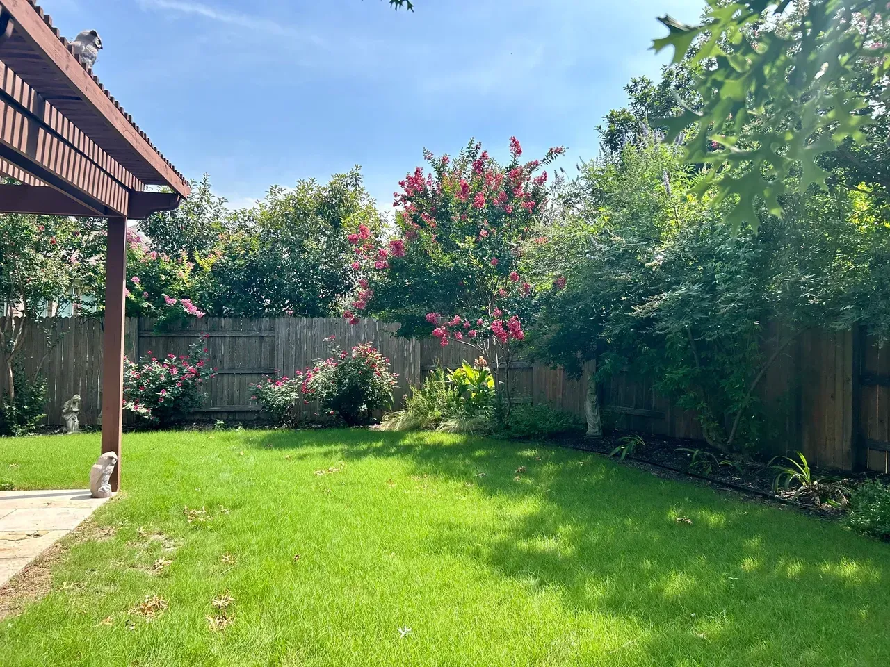 Green backyard with lush grass, flowers, and a wooden fence under a sunny sky.