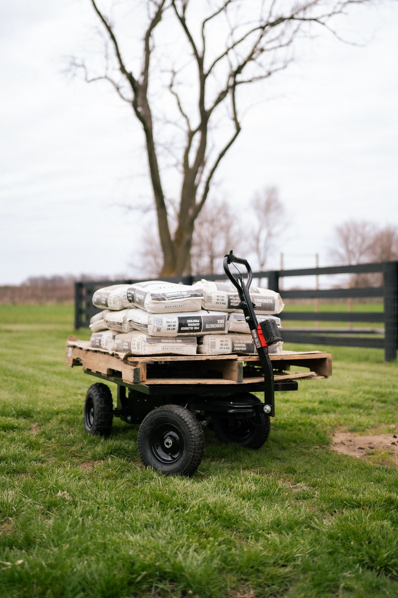 A small, black wheeled cart loaded with bags on a wooden pallet and a shovel, in a grassy field.