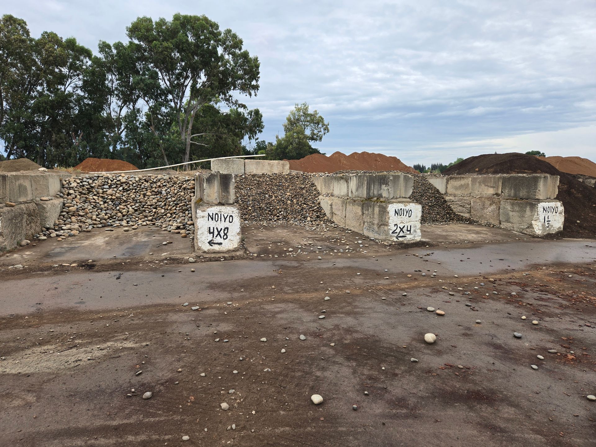 Dry, muddy yard with low stone walls, small white markers, and trees under a cloudy sky.