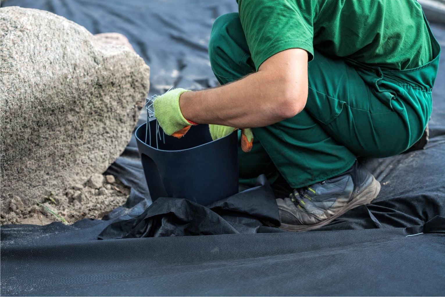 Worker in green uniform kneels by a black bucket, handling material on a black tarp near rocks and water.