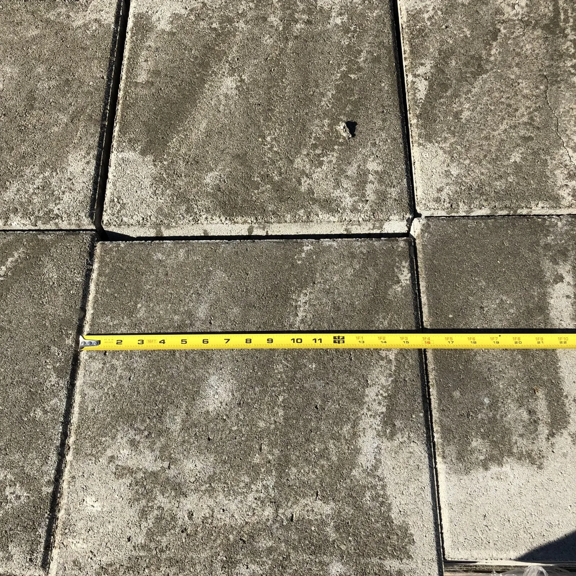 Measuring tape stretched across gray concrete pavers with visible seams and a small crack.