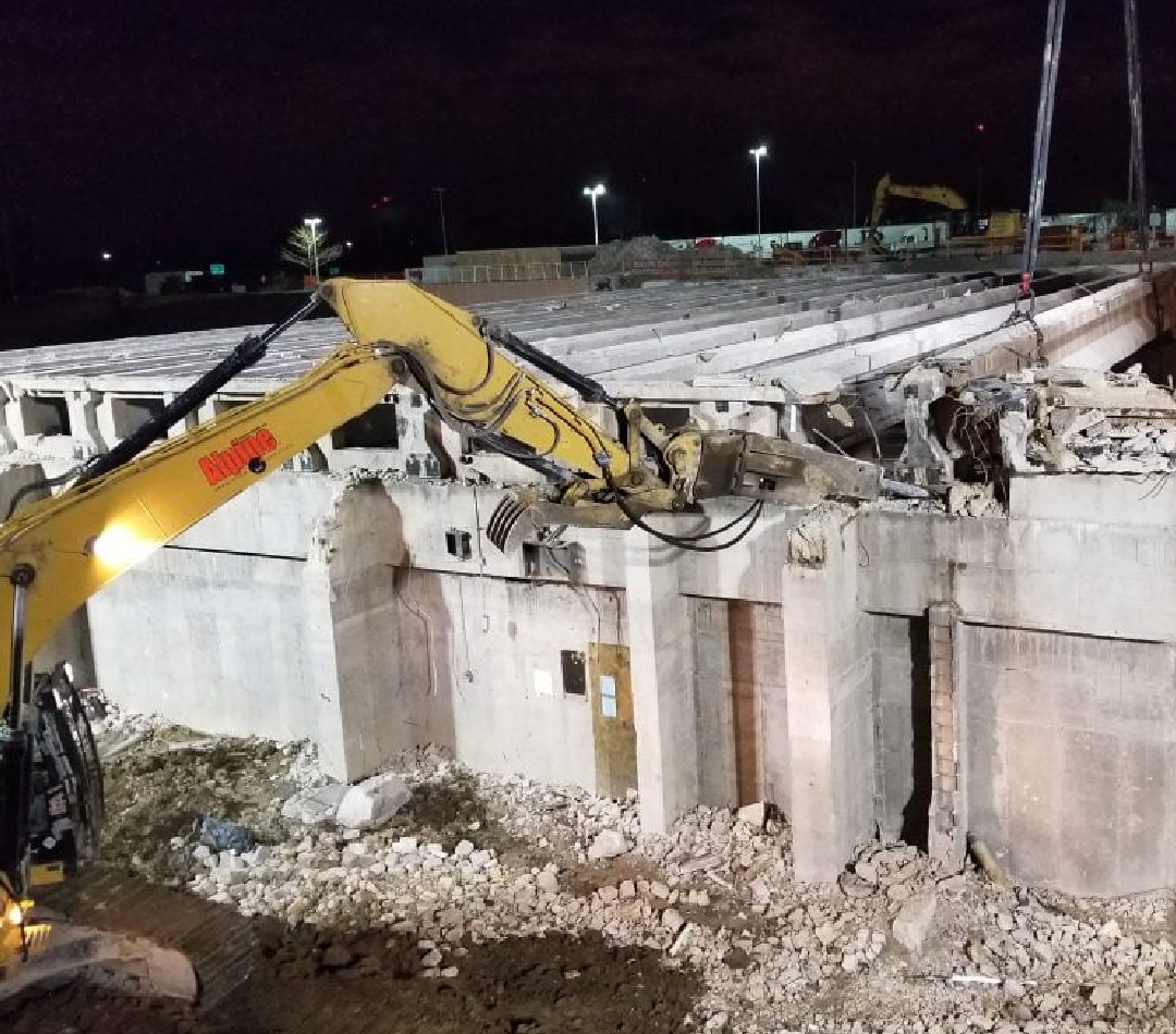A yellow excavator is demolishing a building at night.