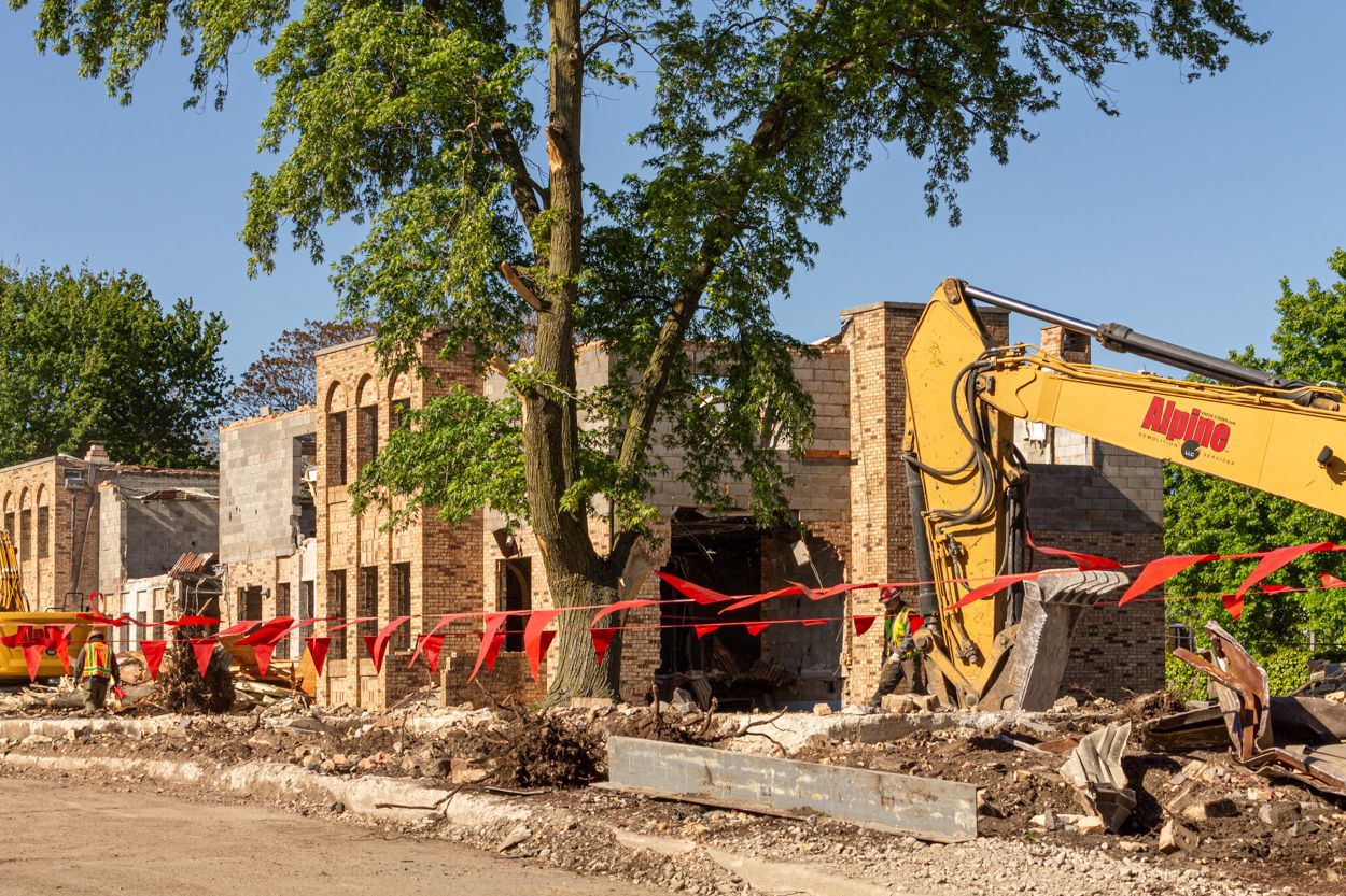 Demolition site with excavator tearing down brick buildings, red caution tape, and a large tree in front