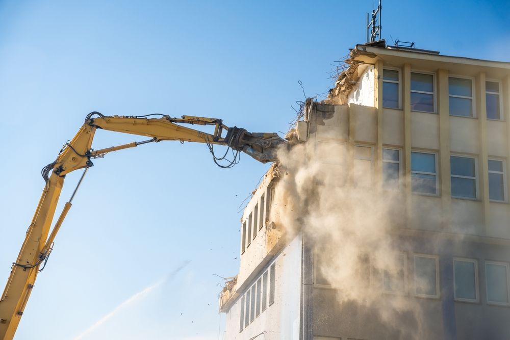 Excavator arm demolishing a building corner, with dust billowing against a blue sky.