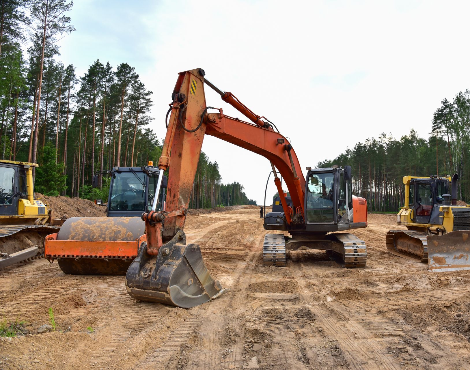 Orange excavator and construction vehicles on a muddy work site with trees in the background