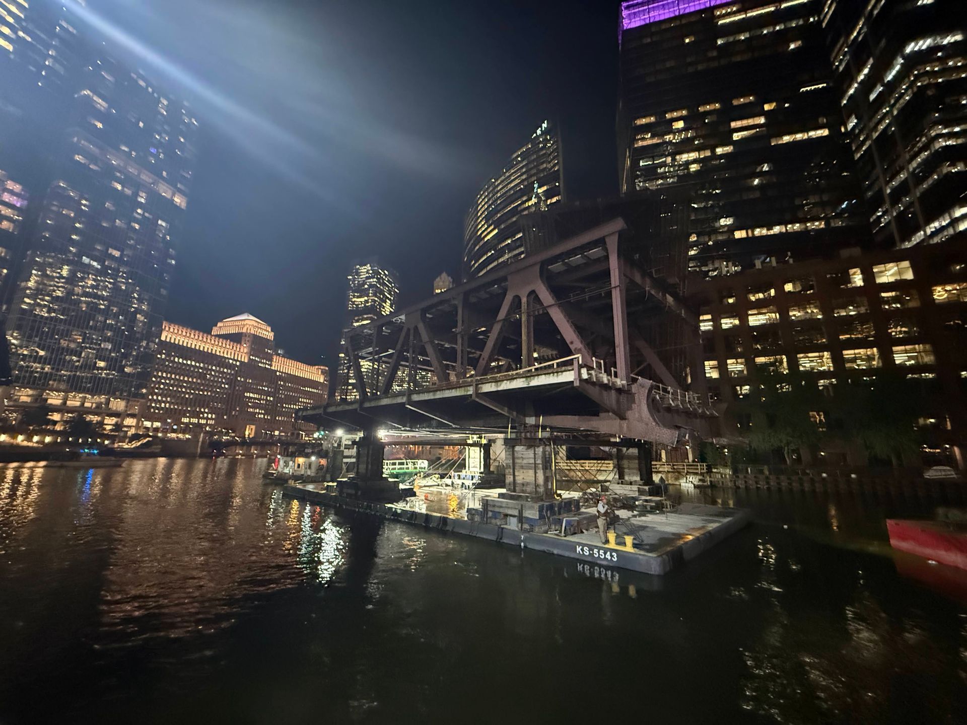 Night city skyline over a river with lit buildings and a steel bridge reflecting on the water.
