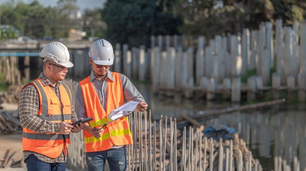 Two construction workers in orange vests and hard hats reviewing plans at a construction site.
