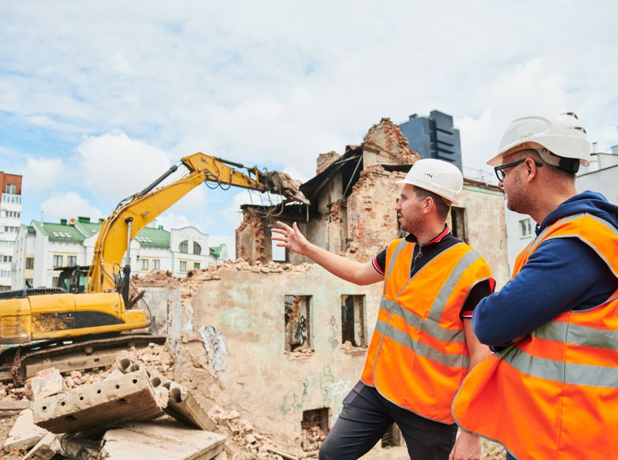 Construction workers in orange vests discuss demolition near an excavator on a rubble-strewn site