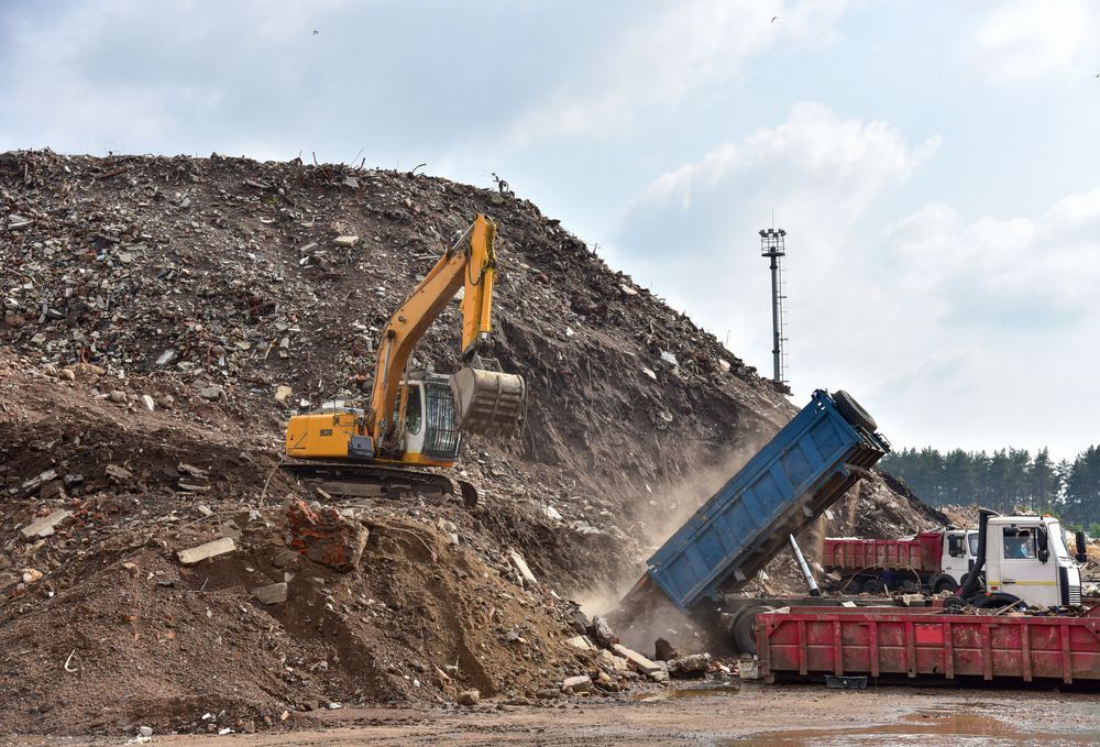 Yellow excavator and blue dump truck unloading soil at a large dirt mound on a construction site.