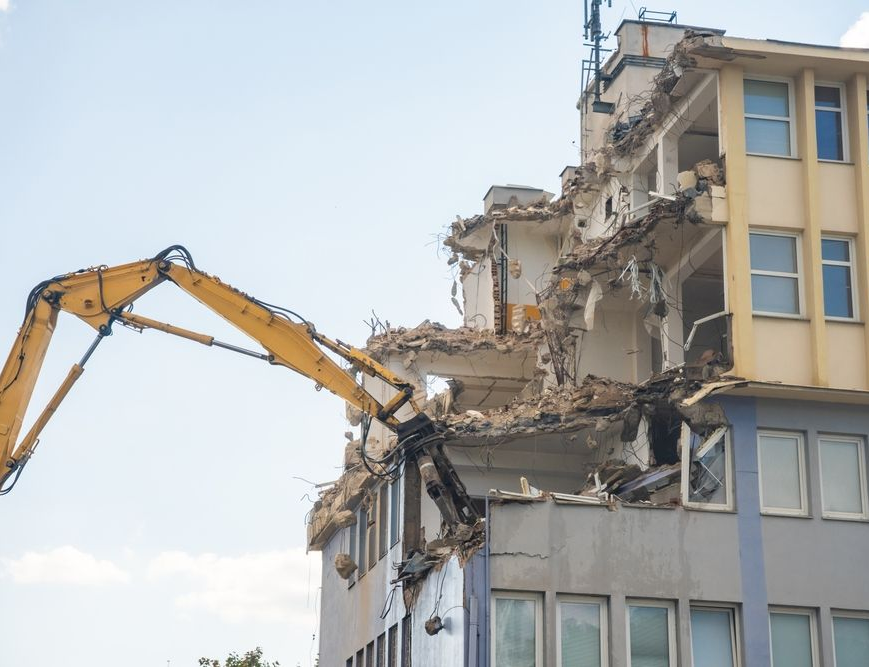 Excavator demolishing the corner of a multi-story building under a blue sky