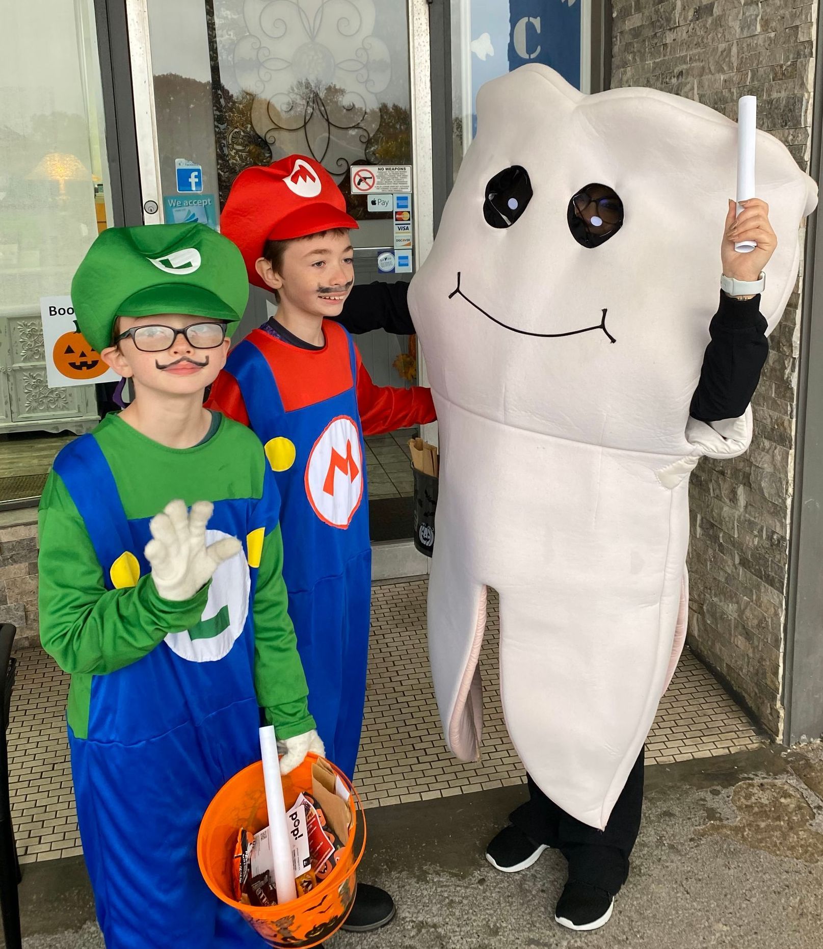 Three children in Halloween costumes posing outside, including Luigi, Mario, and a white ghost-like costume.