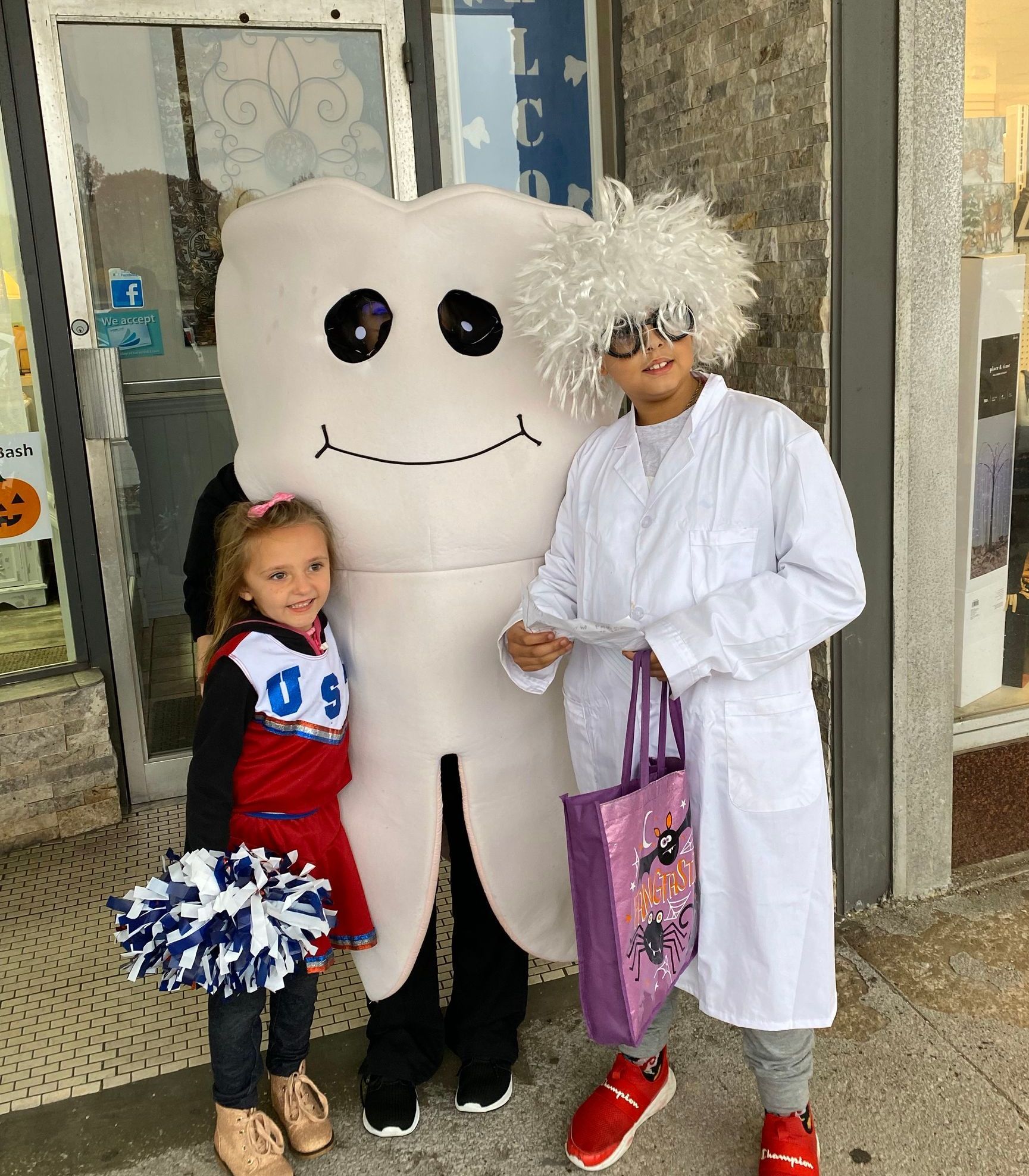 Child with cheerleader pom-poms, a tooth mascot, and a person in a white costume posing at a storefront.