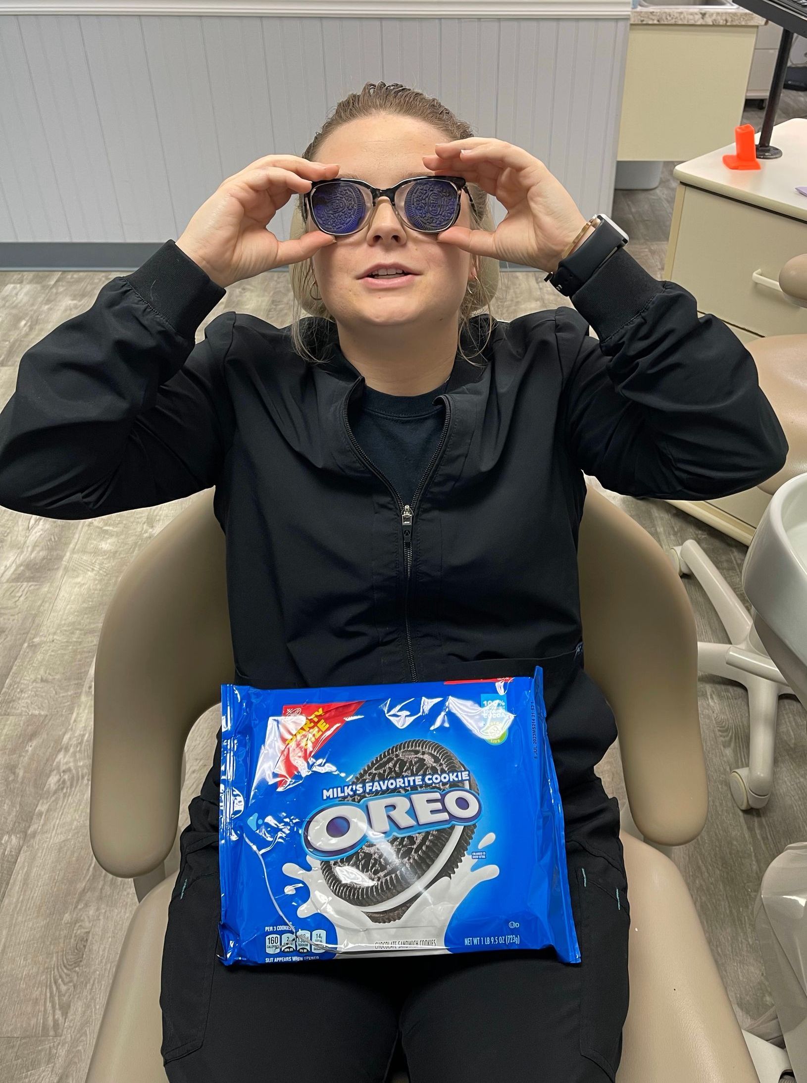 Person seated indoors holding sunglasses to their face, with a blue Oreo package on their lap.