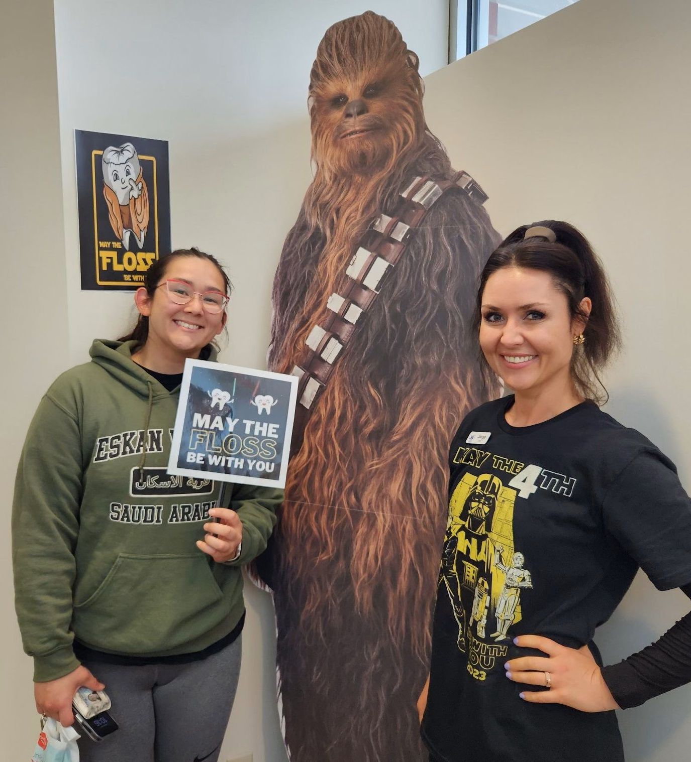 Two people posing with a life-size Chewbacca cutout, one holding a “May the 4th” sign indoors.