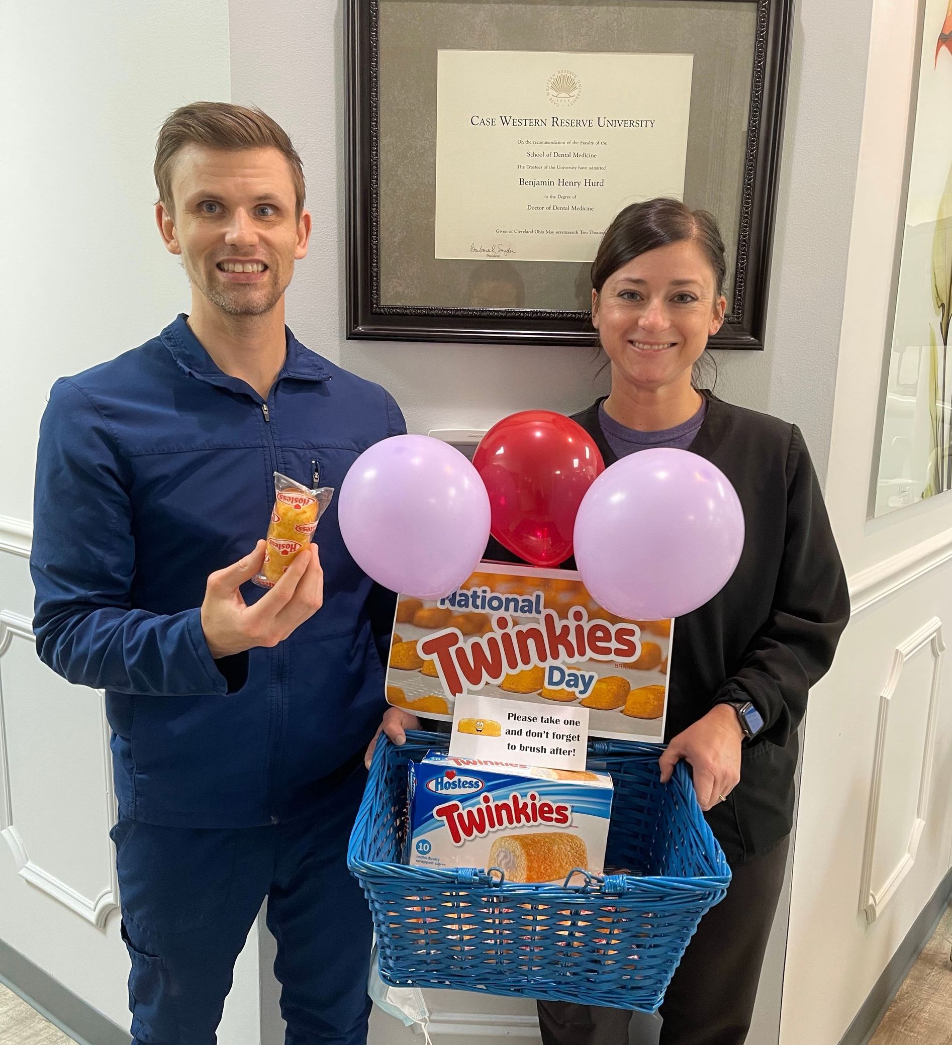 Two people holding a blue gift basket with balloons and snack packs in an office setting