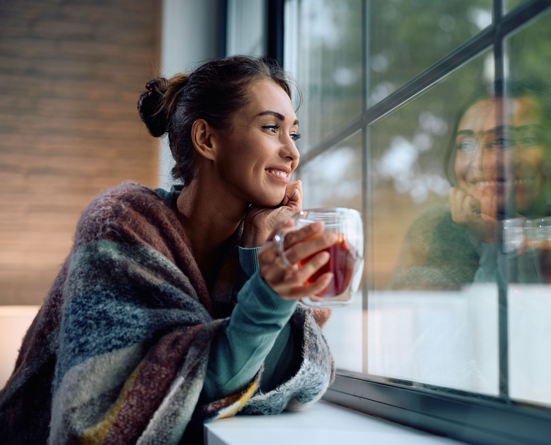 Woman smiling with a mug by a rainy window, looking outside.