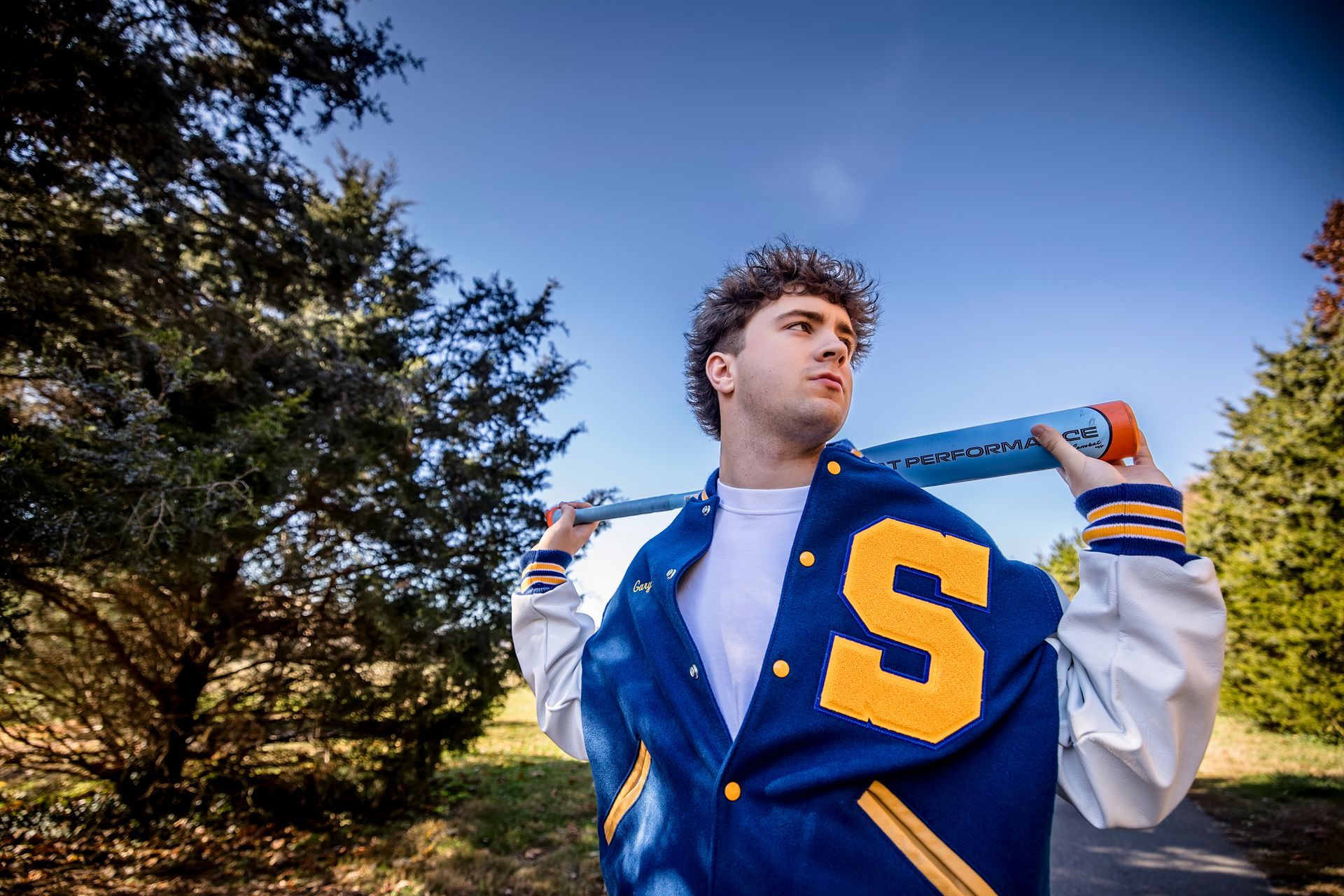 Person in a blue varsity jacket with a yellow “S” stands outdoors, holding it open against a blue sky.