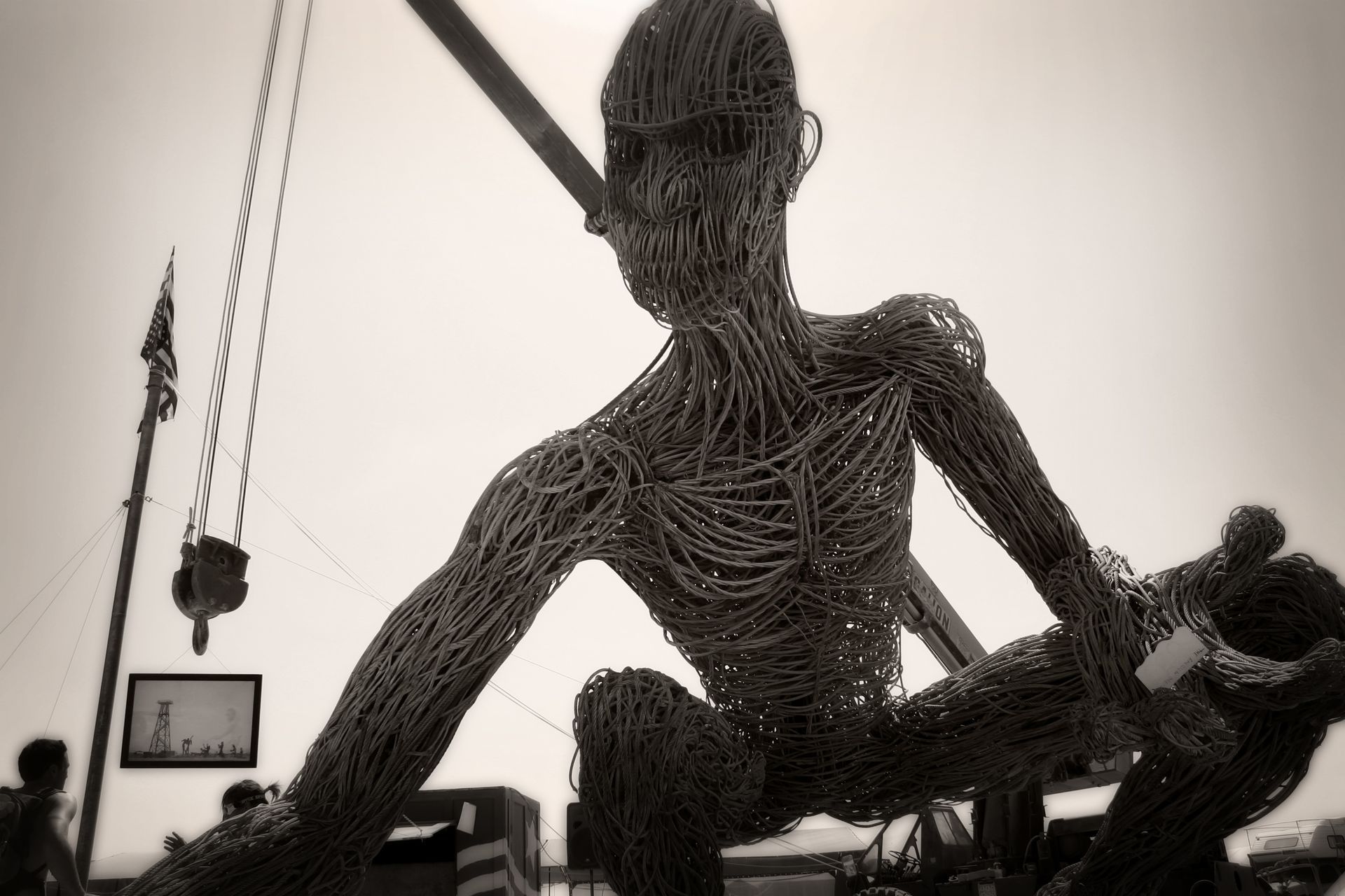 Large woven sculpture of a seated human figure indoors, holding a long object, with hanging art nearby