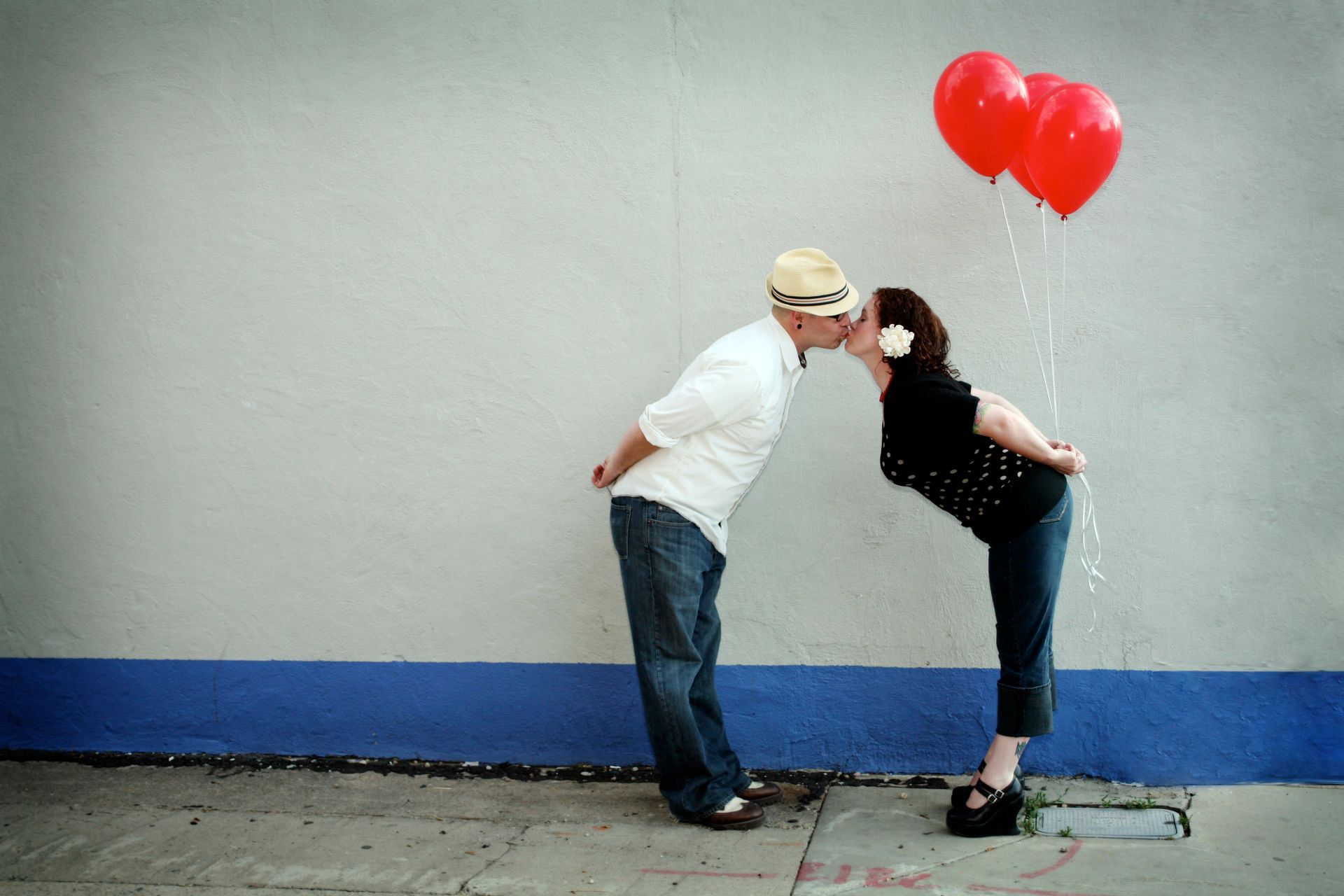Two people leaning toward each other beneath red heart balloons against a gray wall