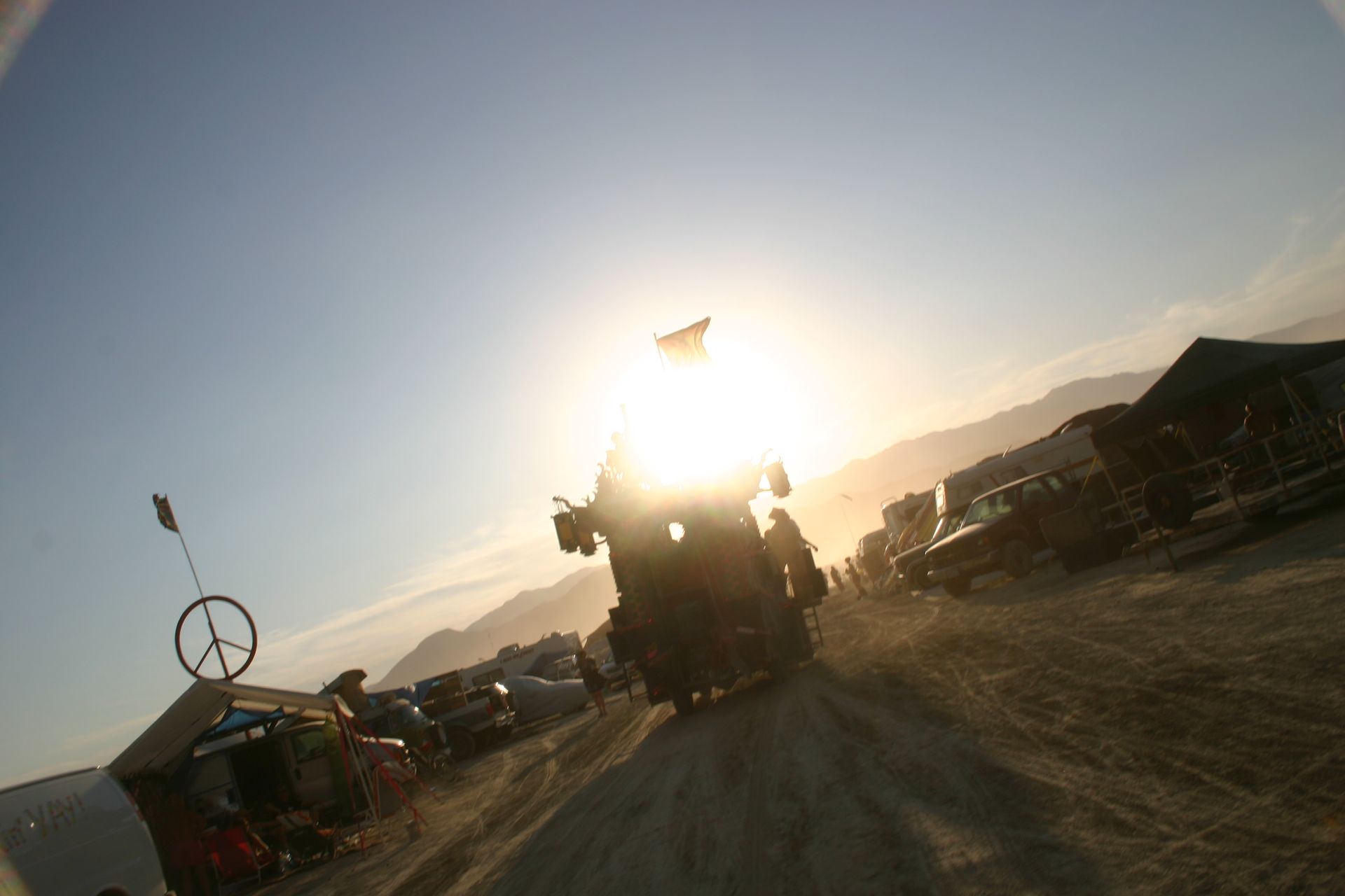 Sunlit dirt road with a large tractor or harvester silhouetted between parked vehicles at sunset