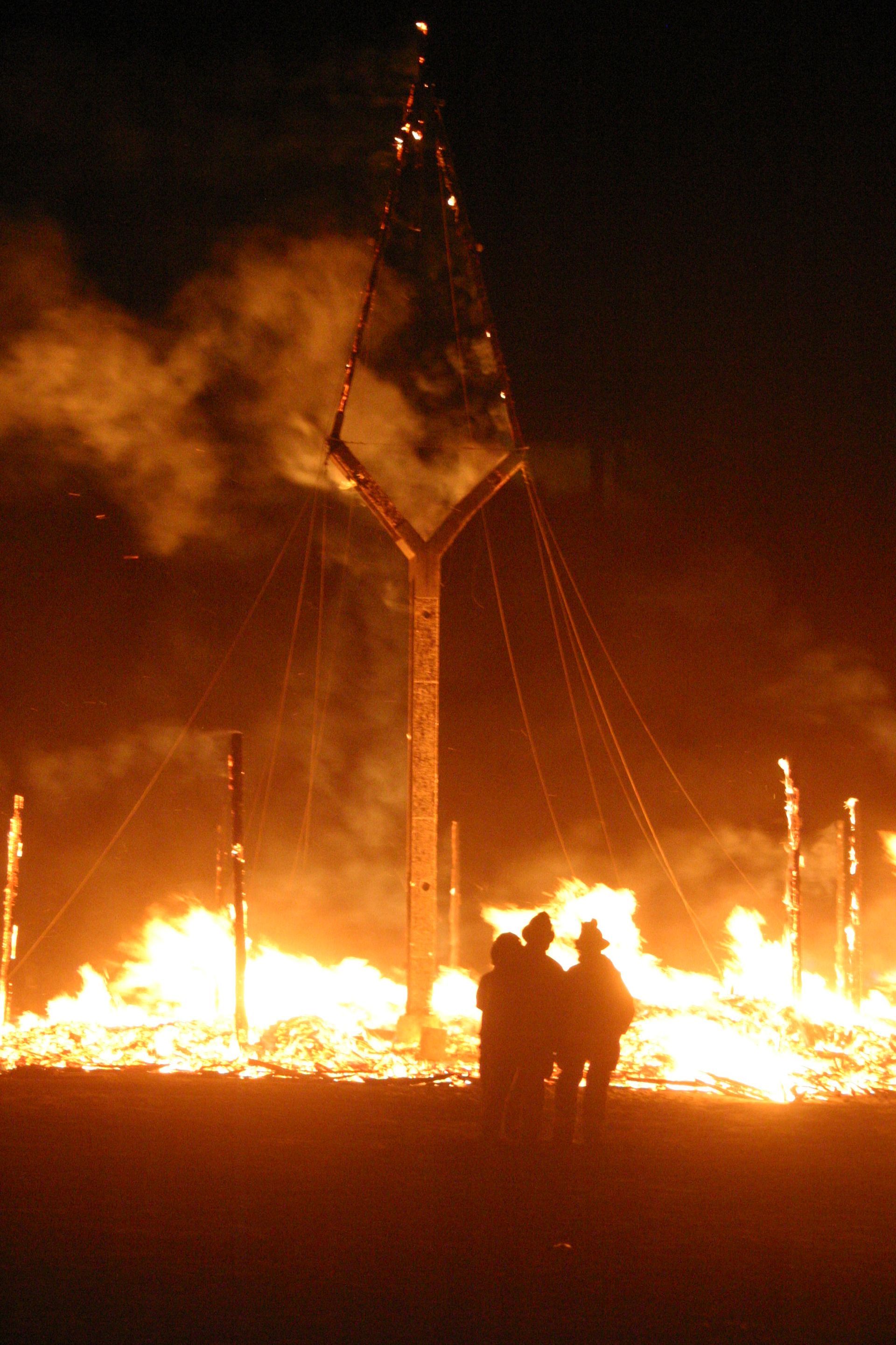 Nighttime fire engulfs a structure, with two silhouetted figures standing in the foreground