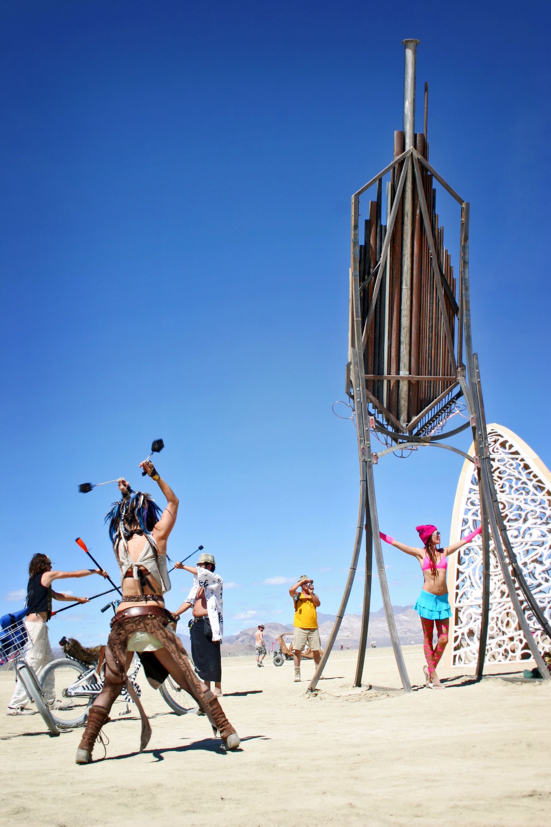 People performing a dance around a tall wooden sculpture on a sandy beach under a clear blue sky