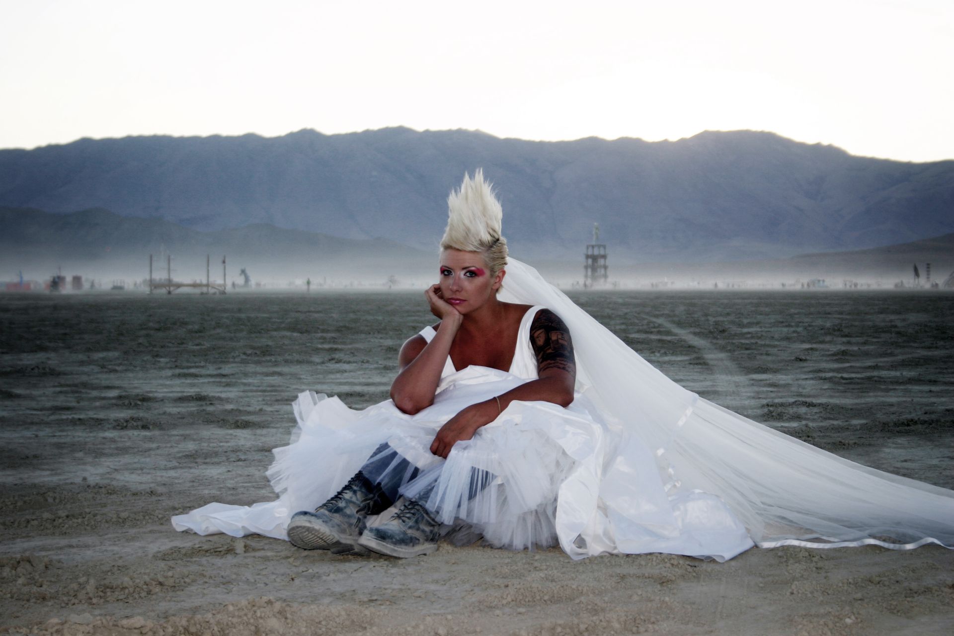 Person in a white outfit and veil sits on a desert plain with mountains in the background.