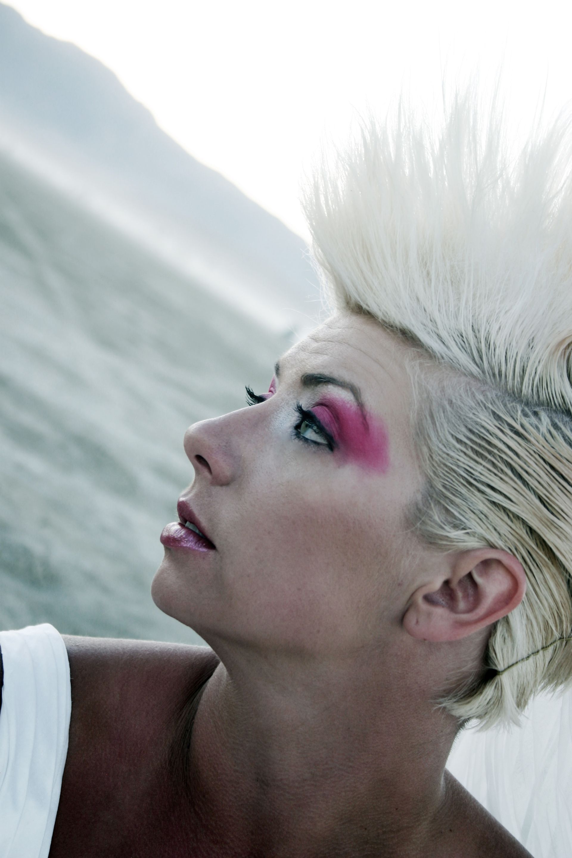 Person with platinum spiked hair and pink eye makeup gazes upward in profile against a soft mountain backdrop