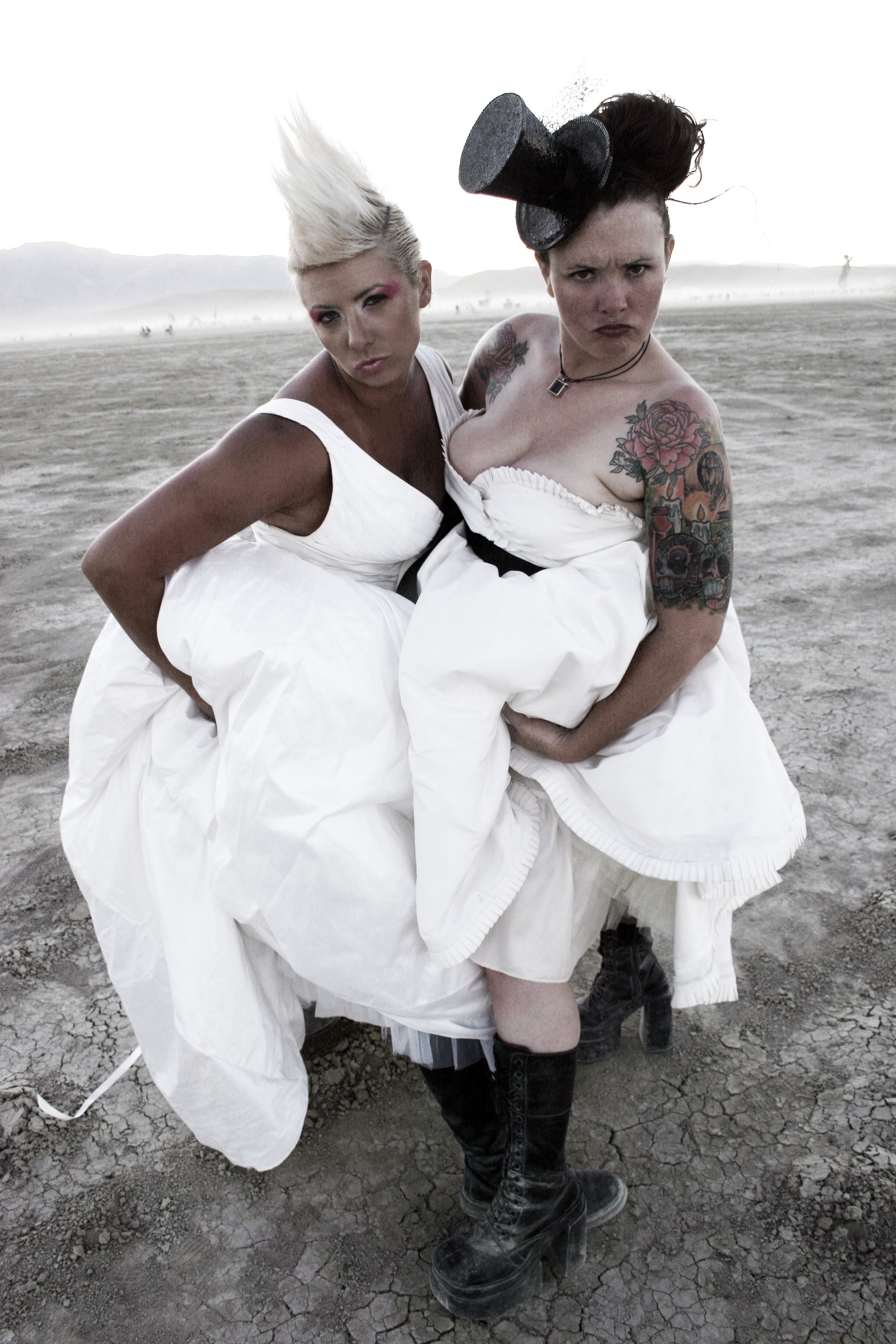 Two people in white dresses stand in a barren field, posing with dramatic hairstyles and black boots.