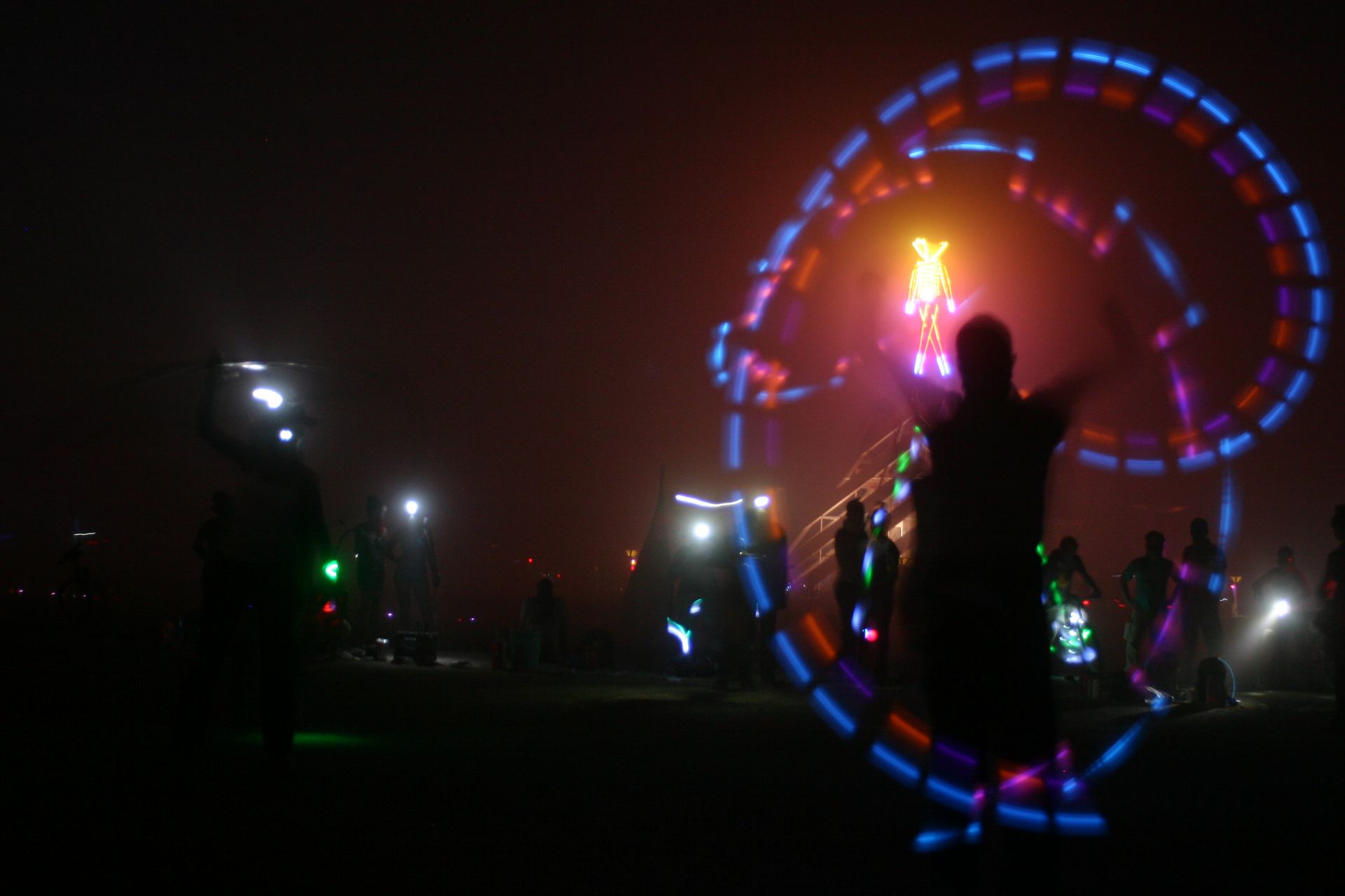Silhouetted performer in dark field, spinning glowing blue and red light trails at night.