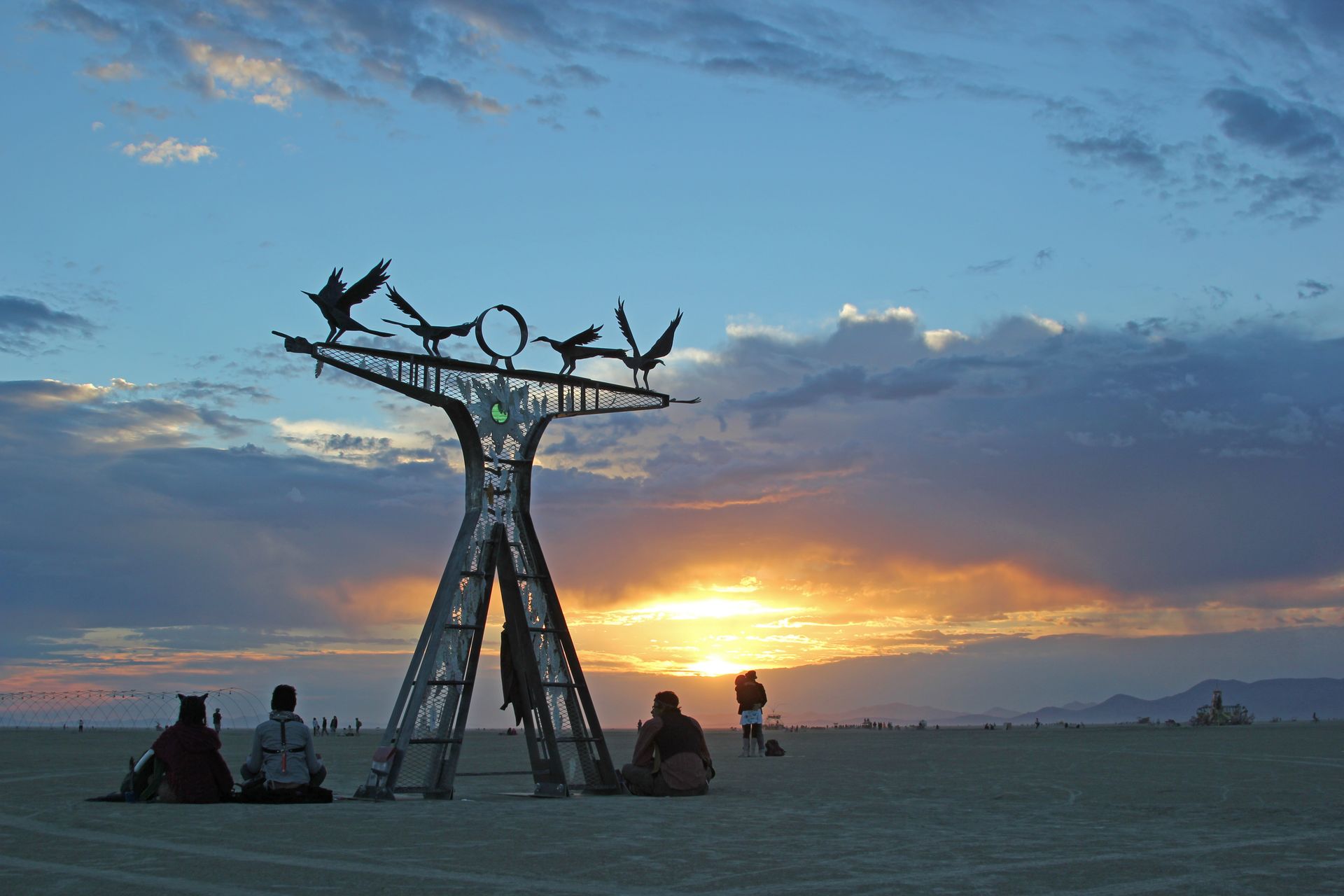 Silhouette of a sculpture and seated people on a beach at sunset, with golden sky and clouds