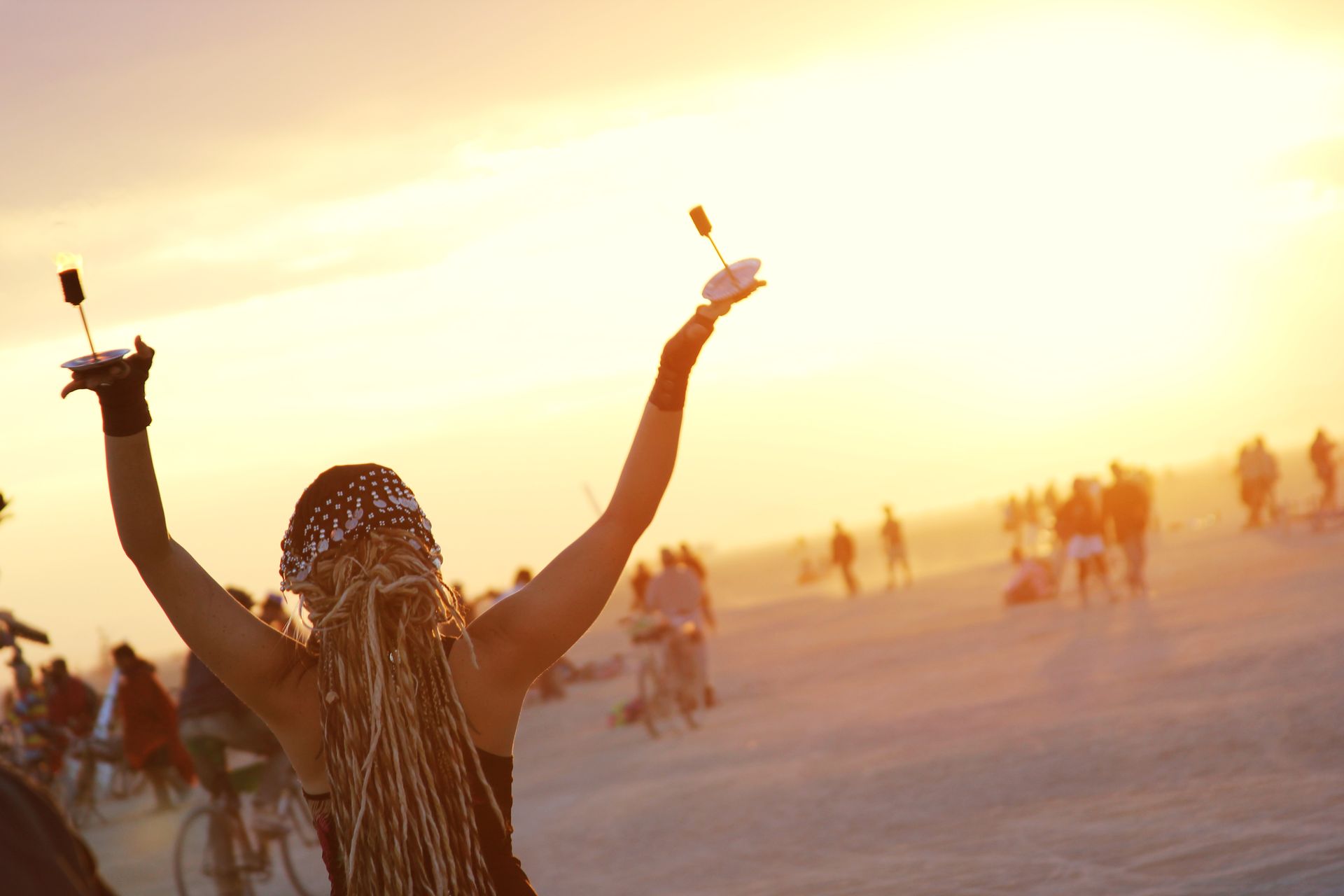 Person with raised arms holding drinks at a sunset festival, with a warm orange sky and distant crowds