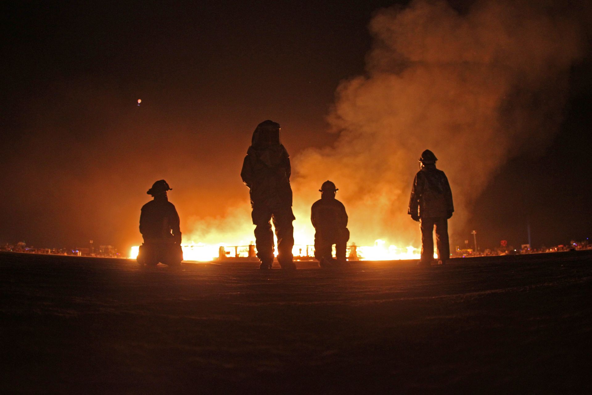Silhouetted people stand before a large fire at night, with smoke rising in the dark.