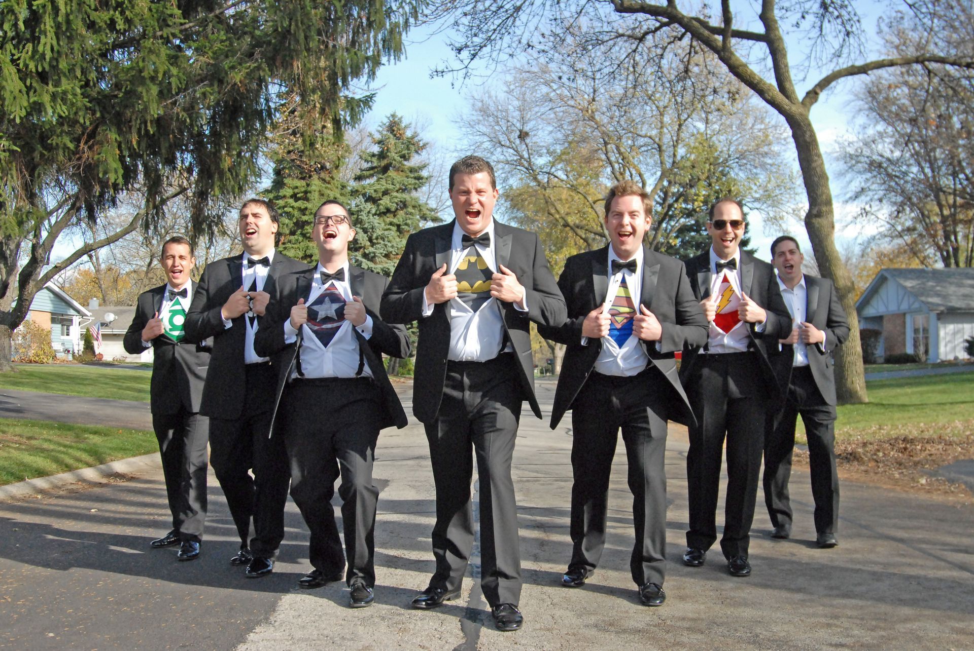 Six men in suits holding drinks and walking outdoors on a sunny path.