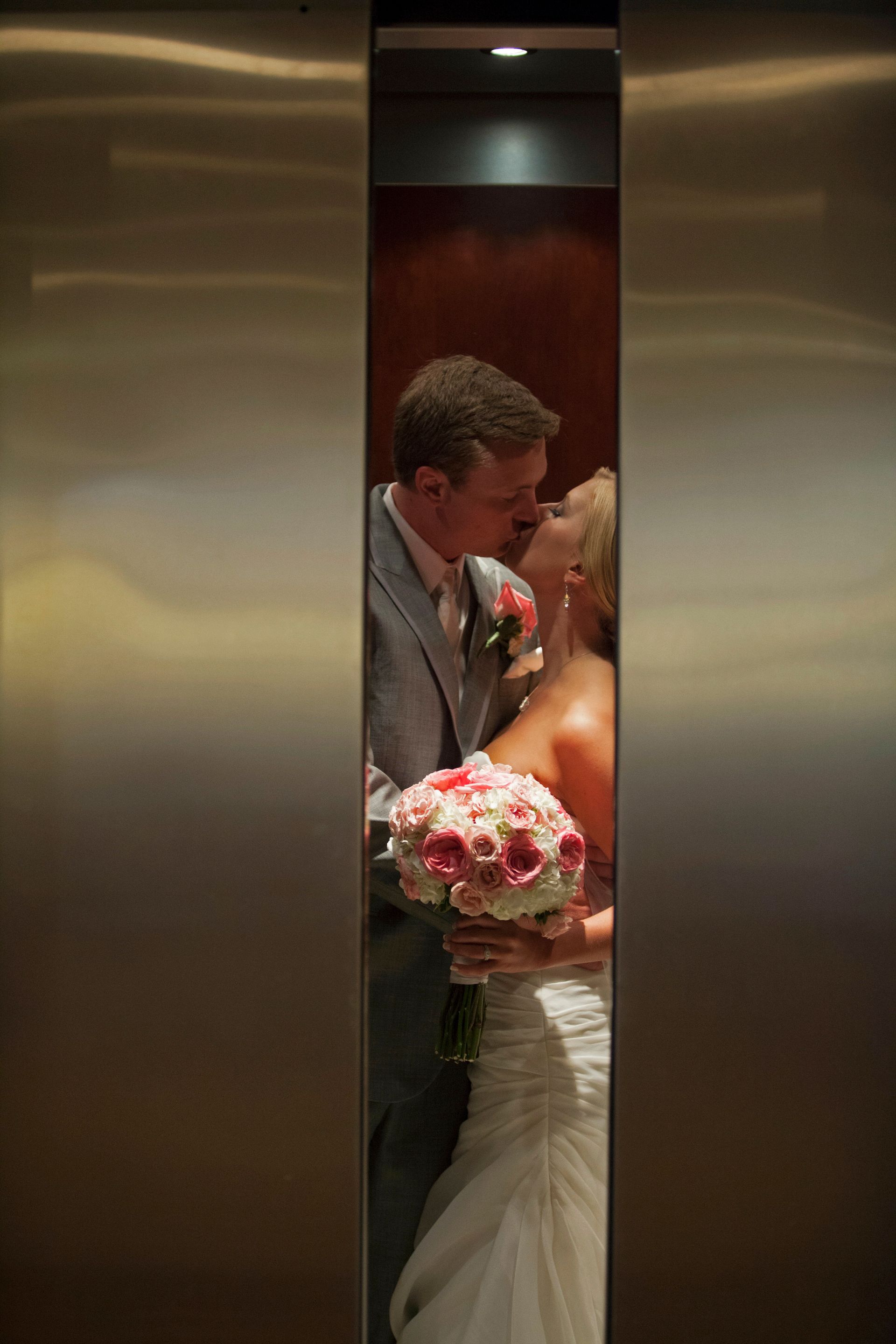 Bride and groom kissing in an elevator, bride holding a pink bouquet