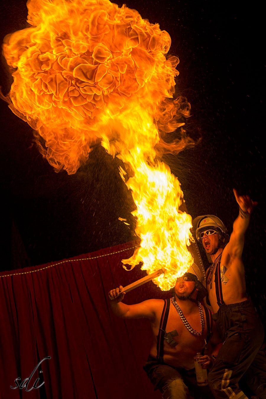 Fire breather exhaling a huge orange flame on a dark stage with red curtains