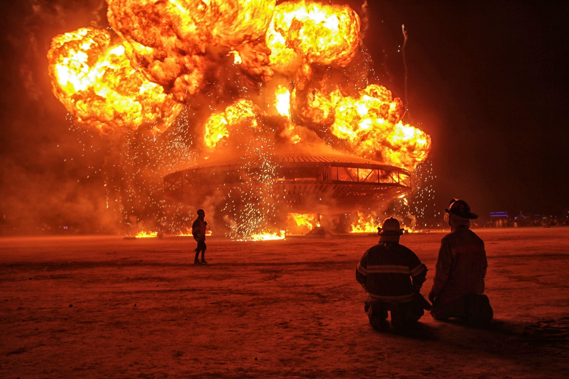 Massive nighttime fire engulfs a building as two seated firefighters watch from the foreground